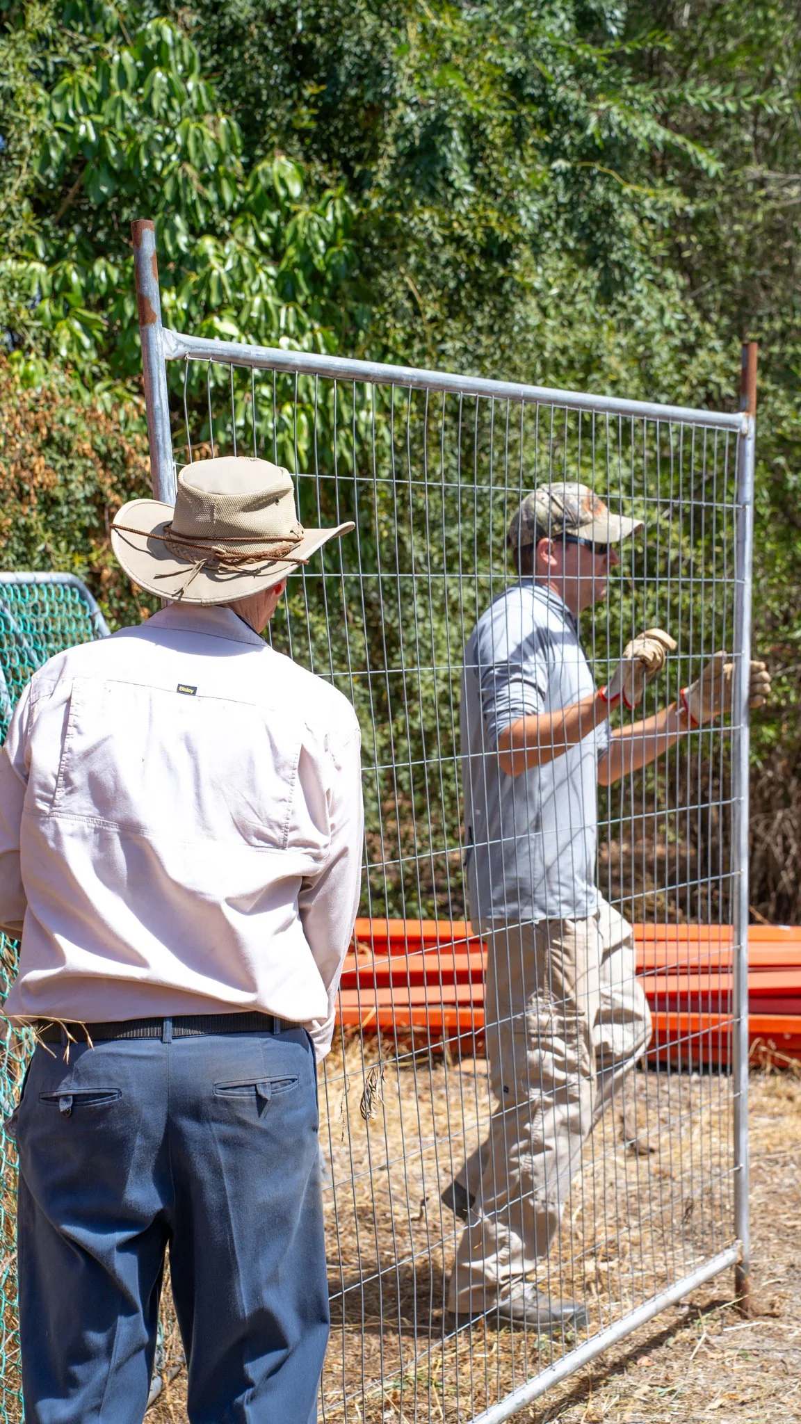 Two men working outdoors with a metal fence, one wearing a wide-brimmed hat and the other wearing a baseball cap, in a wooded area.