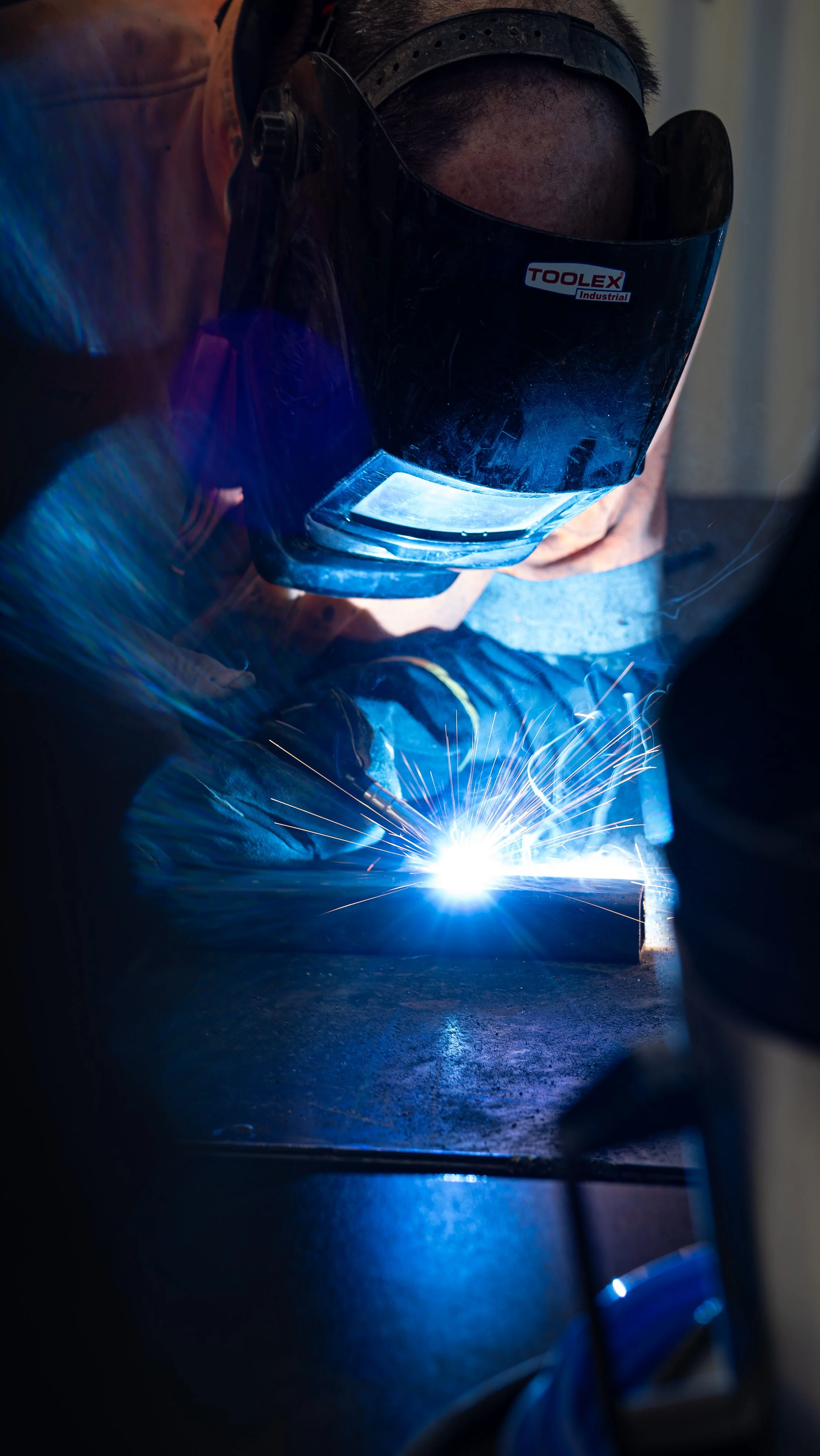 A person is welding metal outdoors, wearing a protective helmet, with a bright arc of light and sparks flying from the welding process.