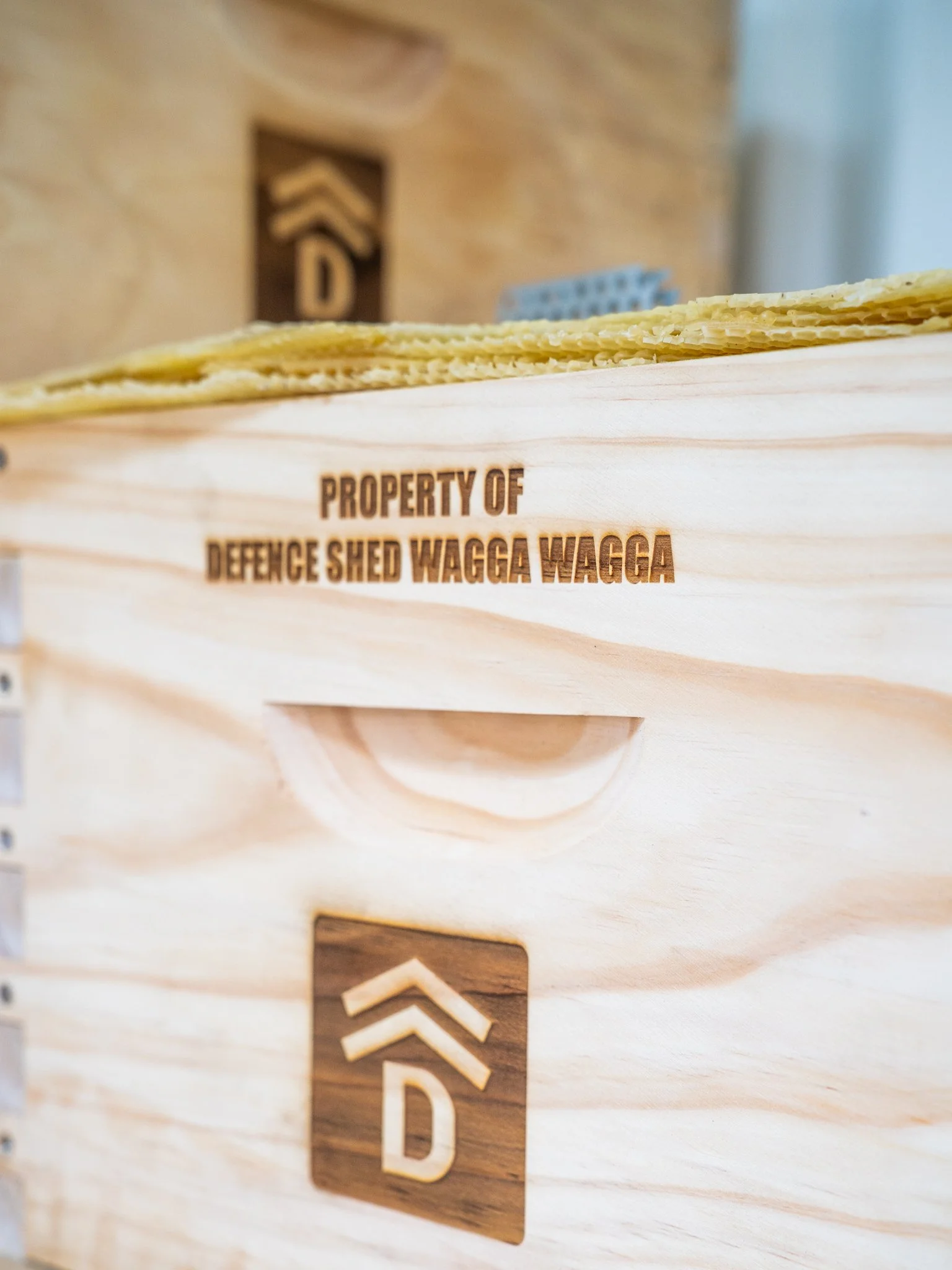 Close-up of a wooden sign with property information, engraved with 'PROPERTY OF DEFENCE SHED WAGGA WAGGA', and a logo or emblem embossed on the surface.