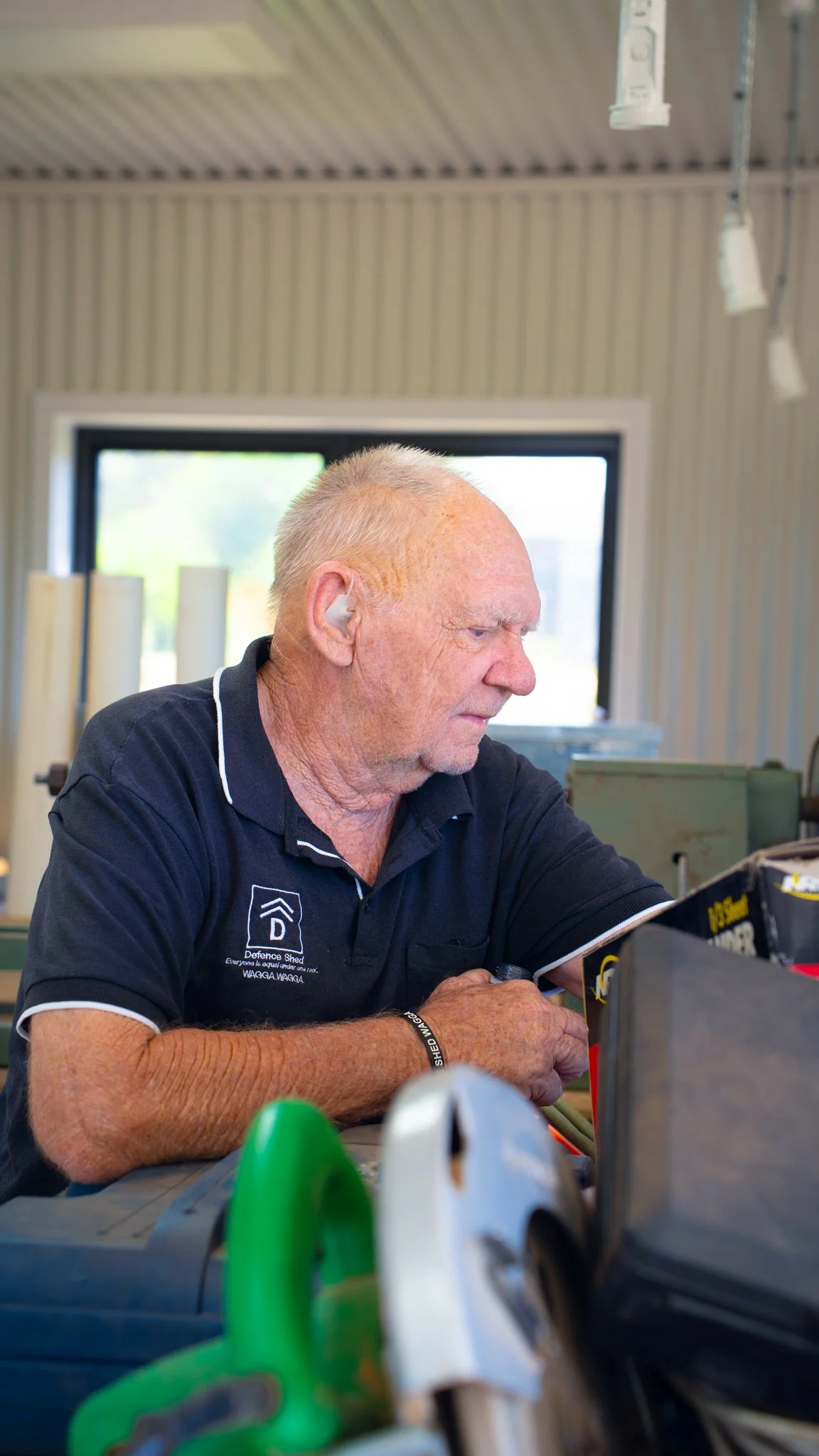An elderly man with short gray hair wearing a black polo shirt working on a project at a workbench inside a workshop. The background shows a window with some sunlight coming through.