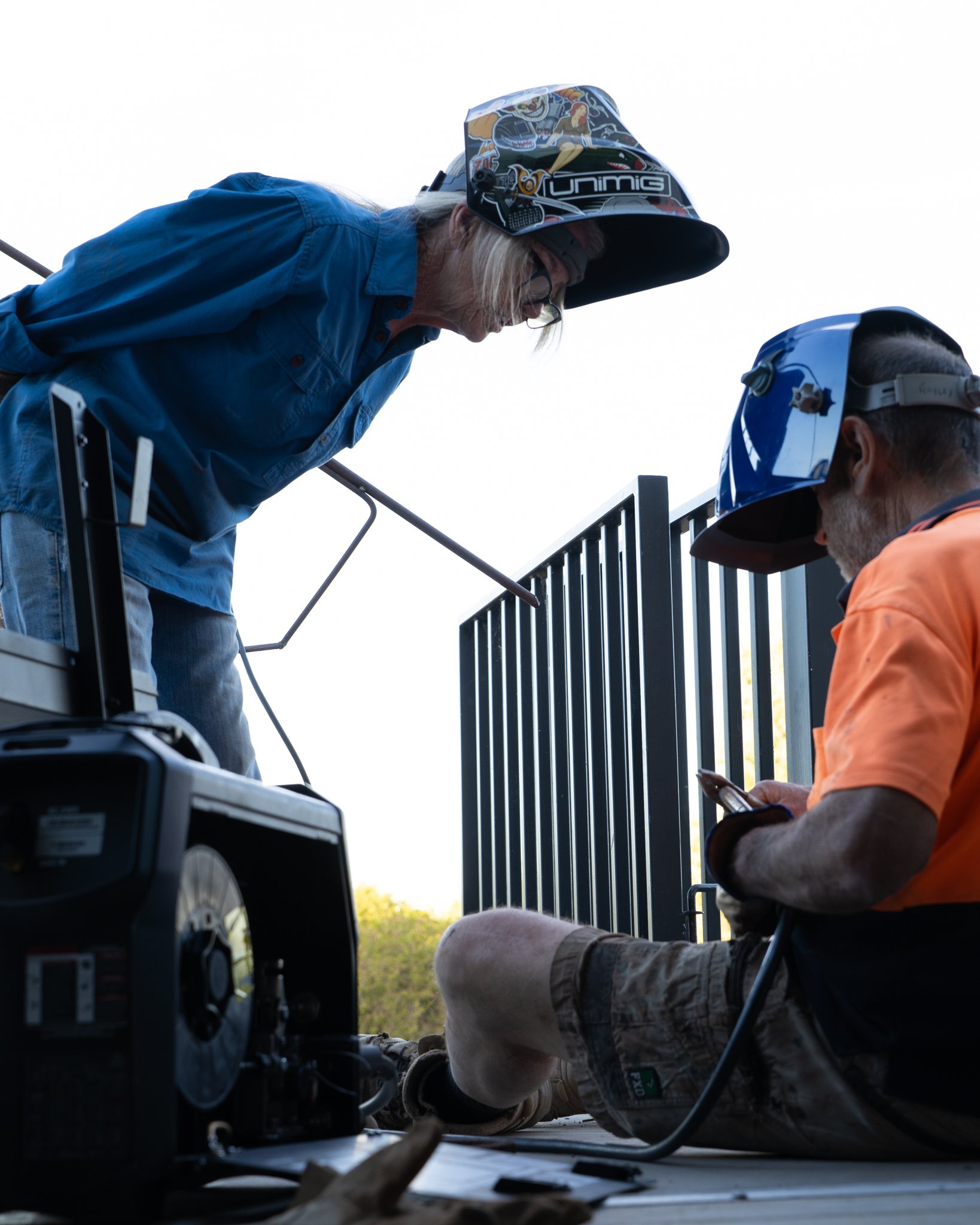 A person lying on the ground underneath a structure, possibly installing or repairing it, with tools and equipment nearby. The individual is wearing a camouflage shirt and beige boots, with a young person in a blue jacket and a helmet assisting.