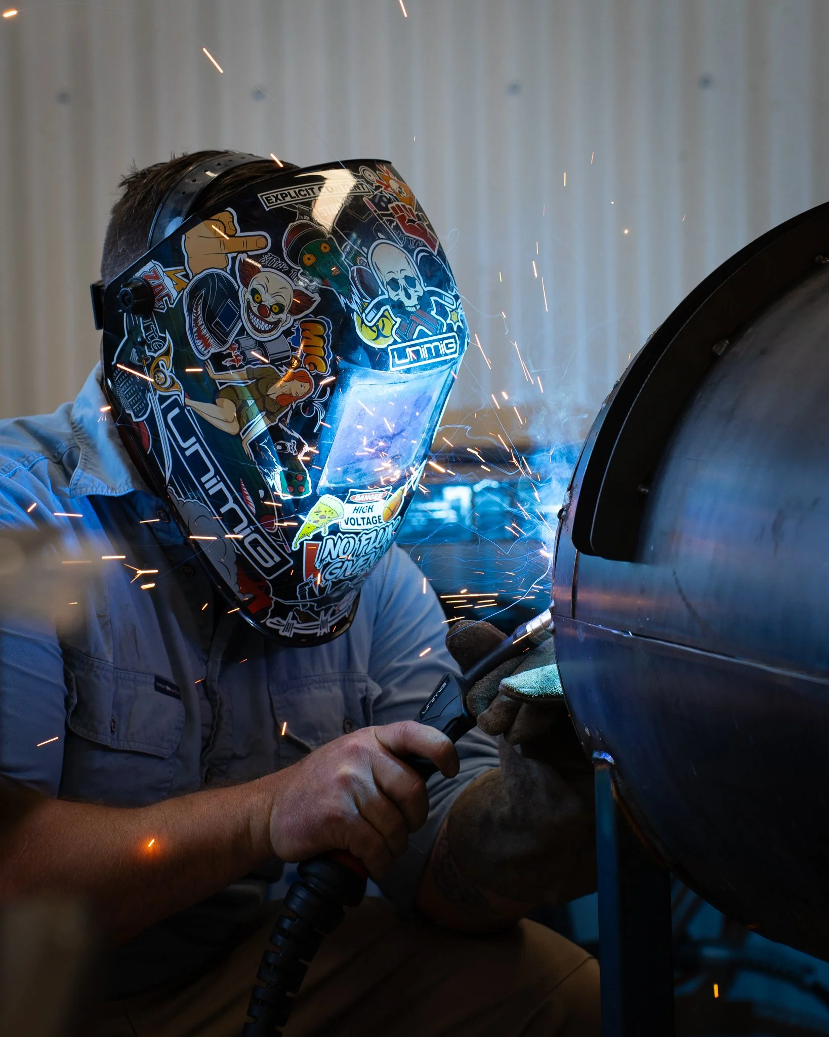 Person welding metal with a welding helmet decorated with stickers. Sparks are flying as the welder works on a metal piece.
