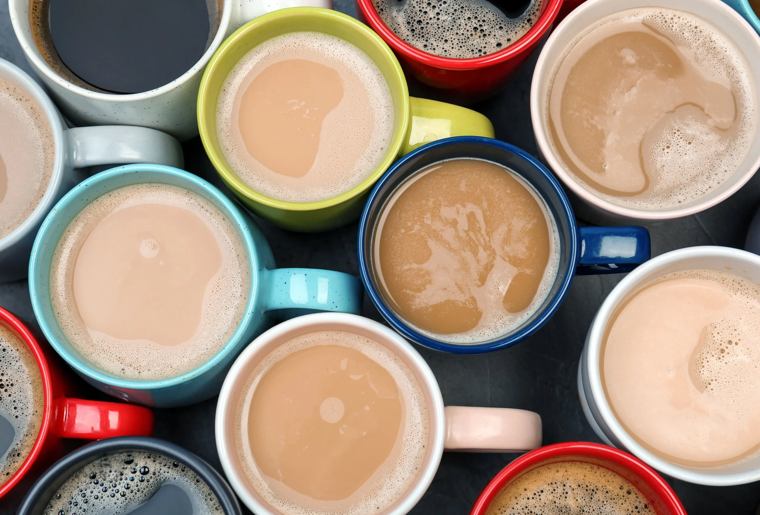 Overhead view of multiple coffee mugs filled with coffee, showing various colors including blue, yellow, white, red, and black on a dark surface.