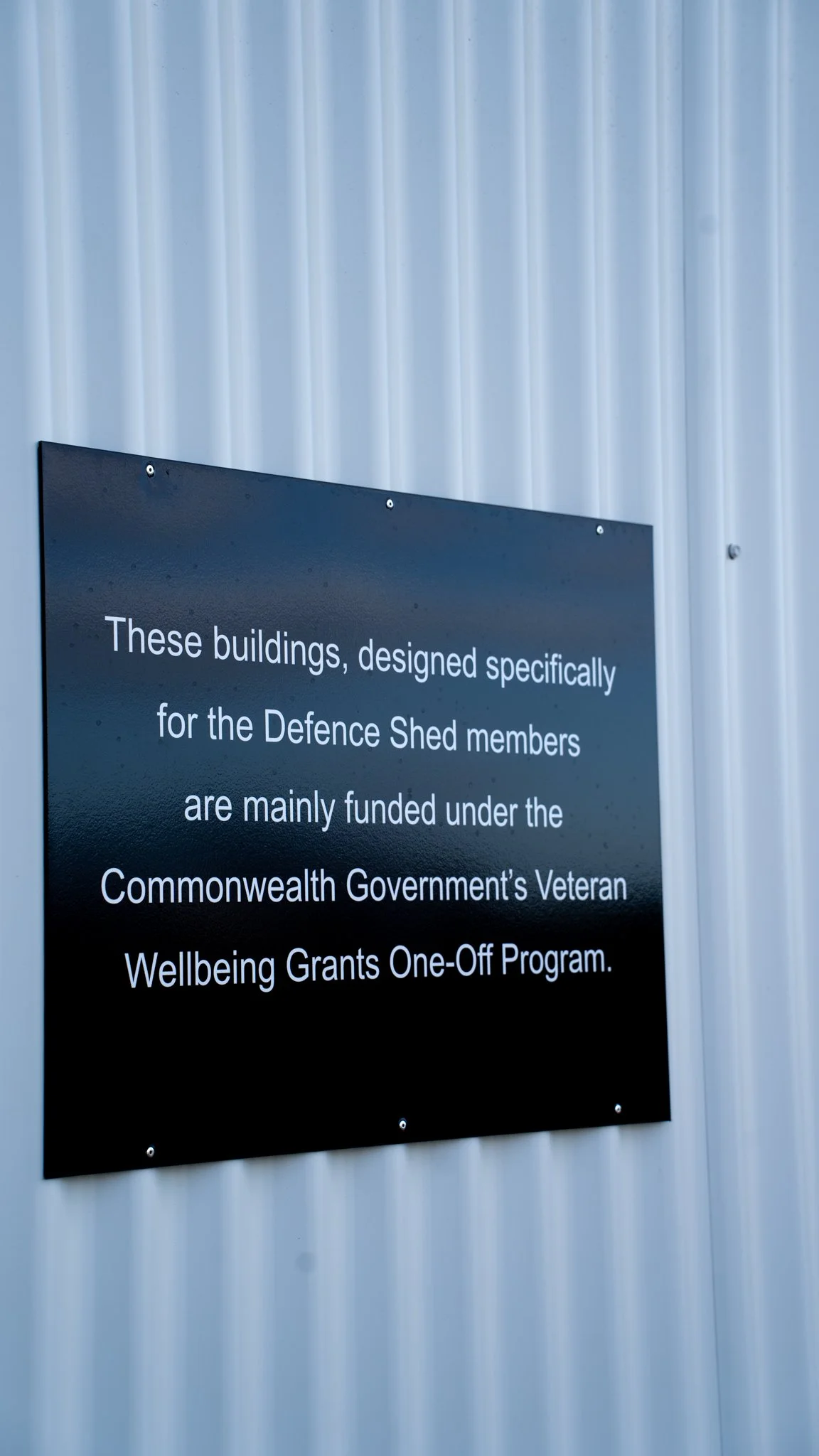 A black sign with white text mounted on a gray metal wall. The sign reads: 'These buildings, designed specifically for the Defence Shed members, are mainly funded under the Commonwealth Government's Veteran Wellbeing Grants One-Off Program.'