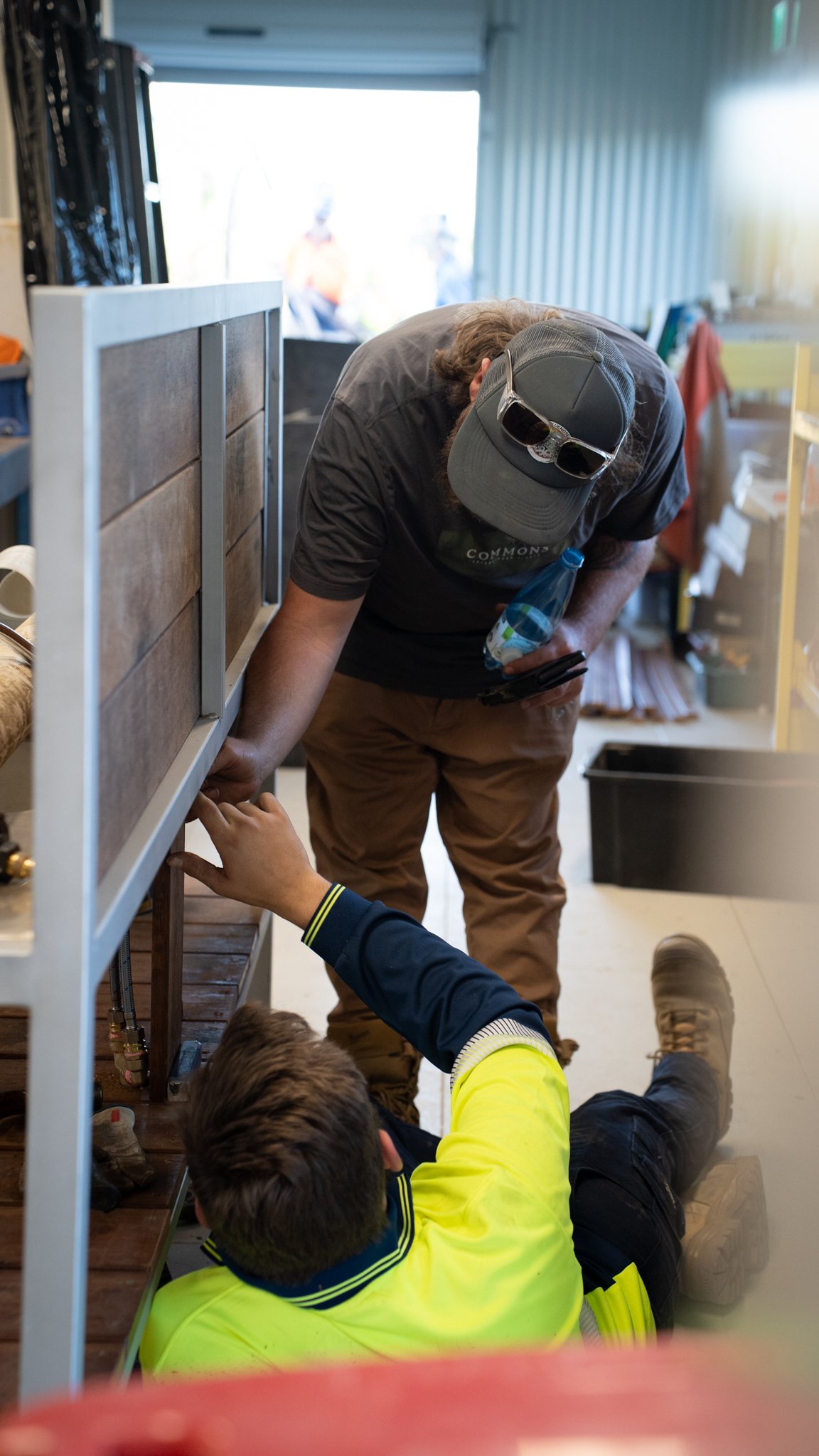 A man and two children working together on a project inside a store or workshop. The man is wearing a cap and glasses on his hat, holding a bottle of water and examining something on the shelf. One child is sitting on the floor, reaching up towards the shelf, while the other child is lying down, wearing a neon yellow jacket.