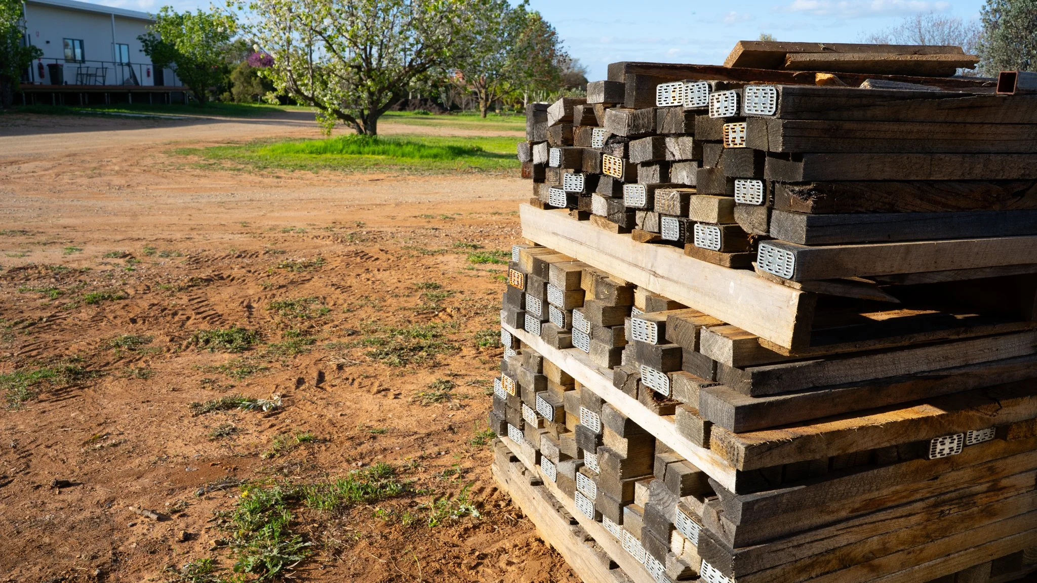 Stack of wooden pallets with metal grid braces on a dirt ground in an open outdoor area, with grass, a tree, and a building in the background.