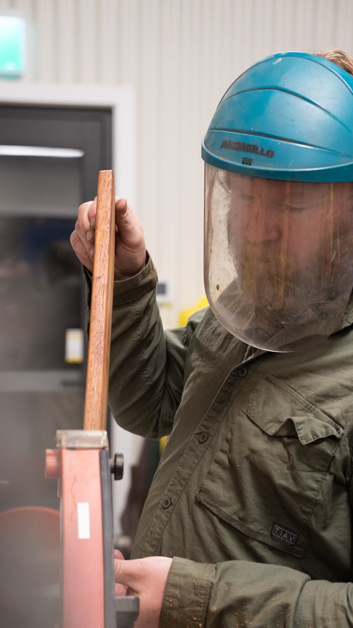 A person working with a machine, wearing a blue helmet with a face shield and a work jacket, holding a wooden object.