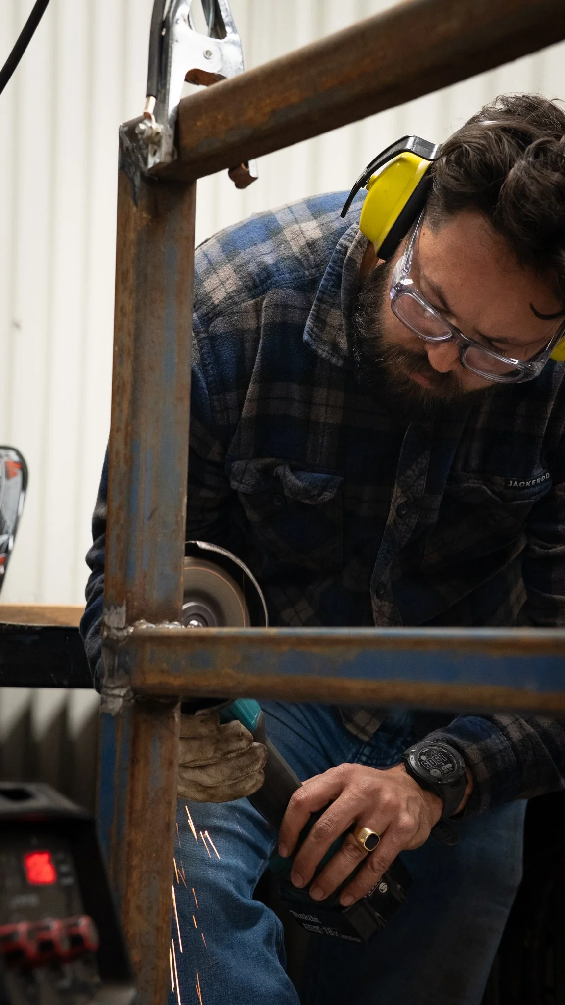 A man welding metal pipes in a workshop, wearing safety glasses, yellow ear protection, and a plaid shirt, with sparks flying from the welding process.