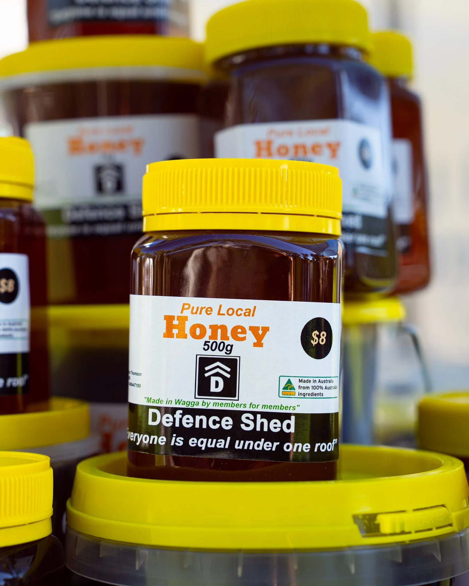 A jar of honey with a yellow lid among other honey jars on a display shelf. The label indicates it is pure local honey, priced at $8, and made in Wagga, Australia, from 100% Australian ingredients.