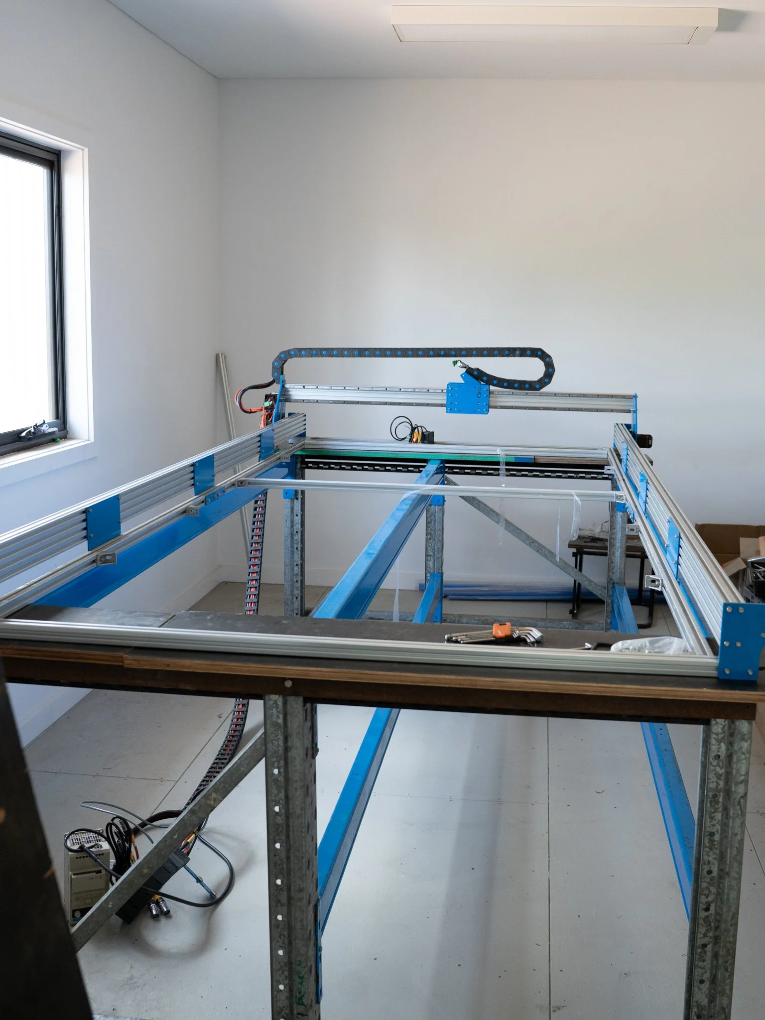 Partially assembled metal frame structure in a bright room with white walls and ceiling, some tools and wires on the floor.