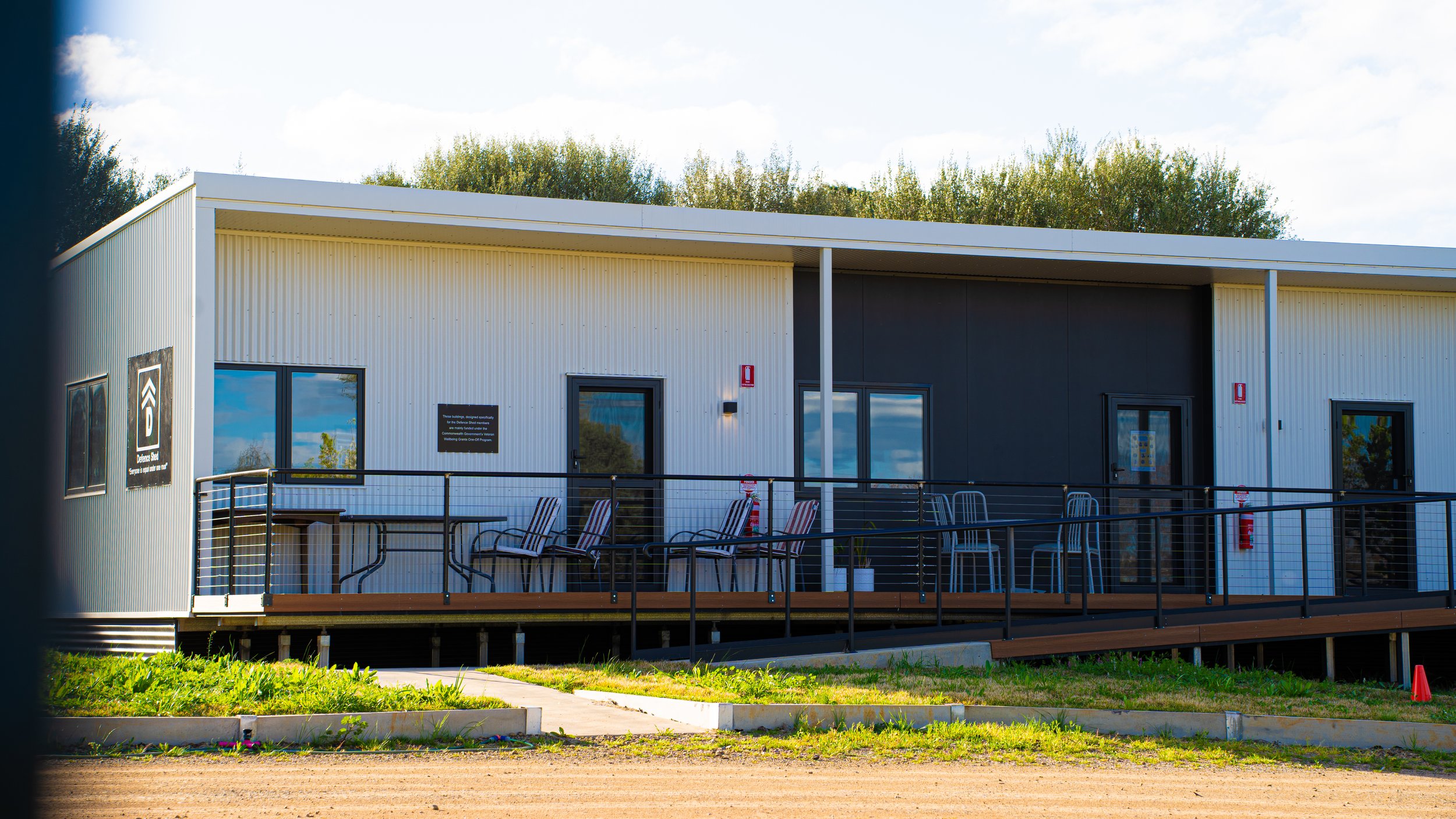 Modern two-story building with metal siding, large windows, and outdoor seating area surrounded by a black railing, featuring a sign indicating it is a Darken Street facility with trees in the background.