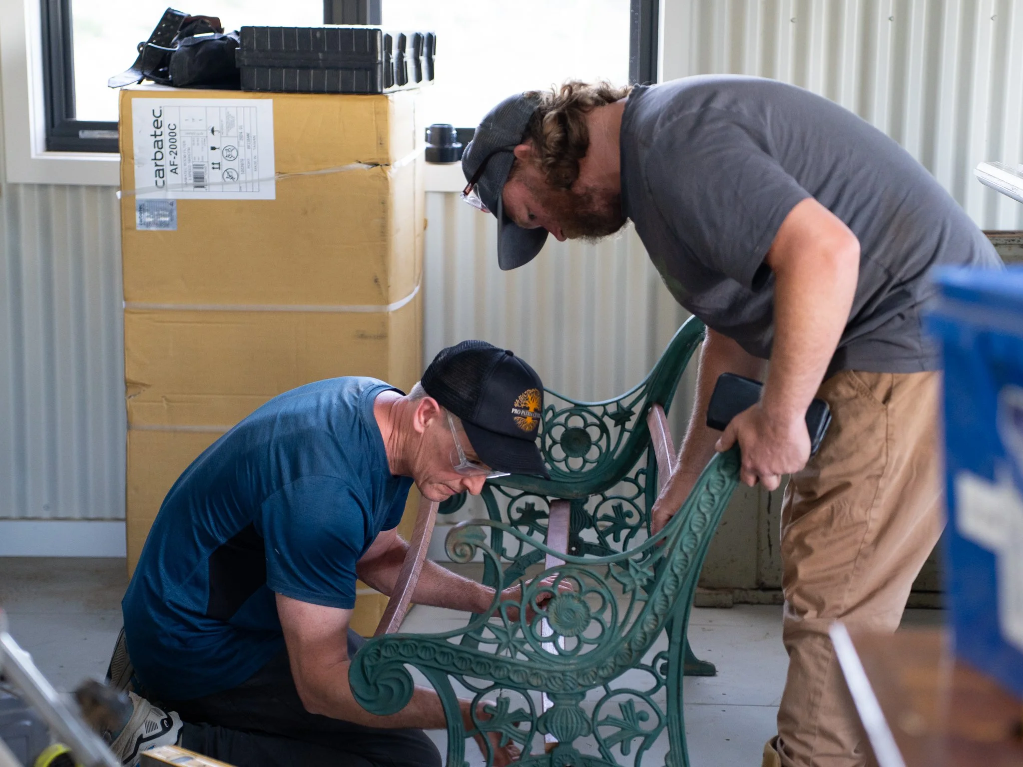 Two men working together on a green metal bench, one kneeling and the other bending over, both wearing safety glasses. A large cardboard box, a black tool case, and other objects are on a table nearby, in a well-lit room with vertical beige paneling.