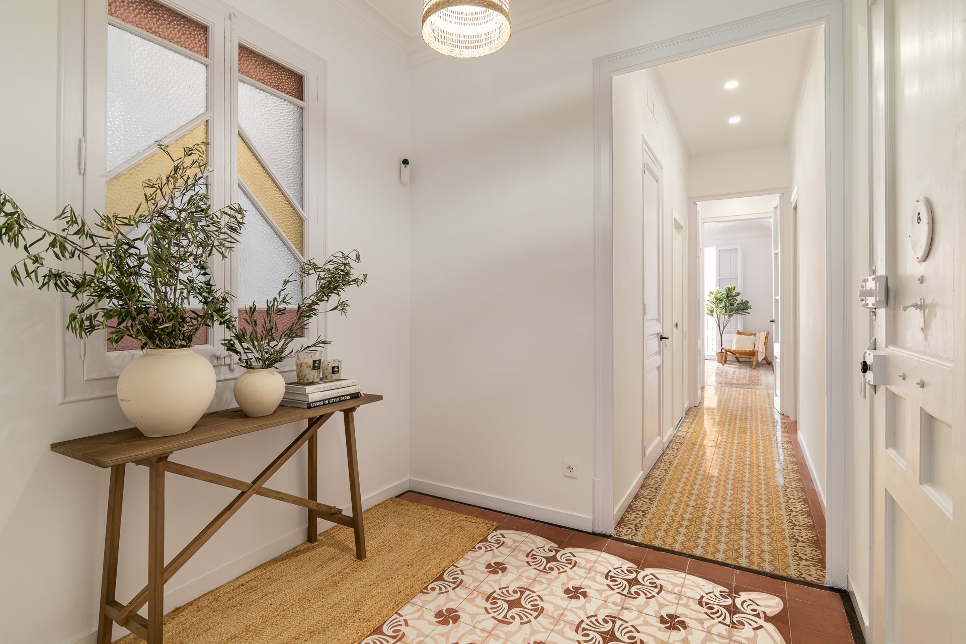 Entrada de casa con paredes blancas, ventanas con cristales decorativos, plantas en jarrones blancos, y pasillo con puertas blancas y piso de azulejos stylish.
