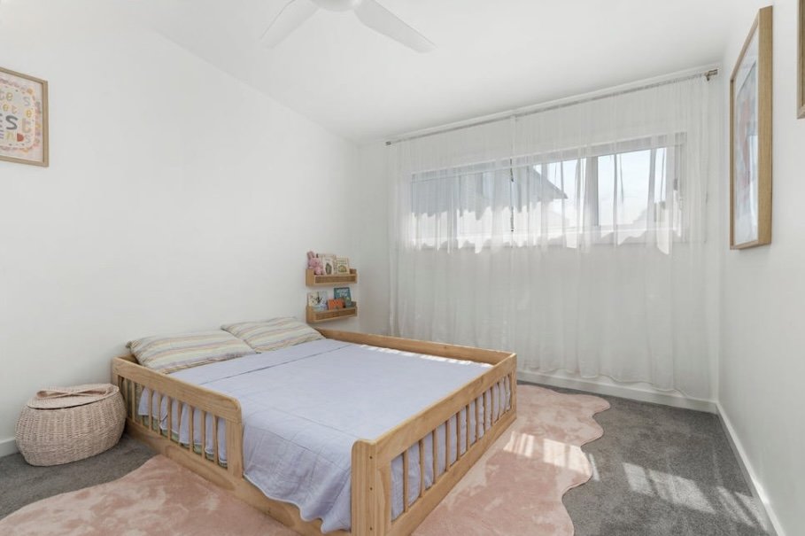 A bedroom with a wooden bed frame, two pillows, a small wall shelf with books, a wicker basket, pink rug, and a large window with sheer curtains.