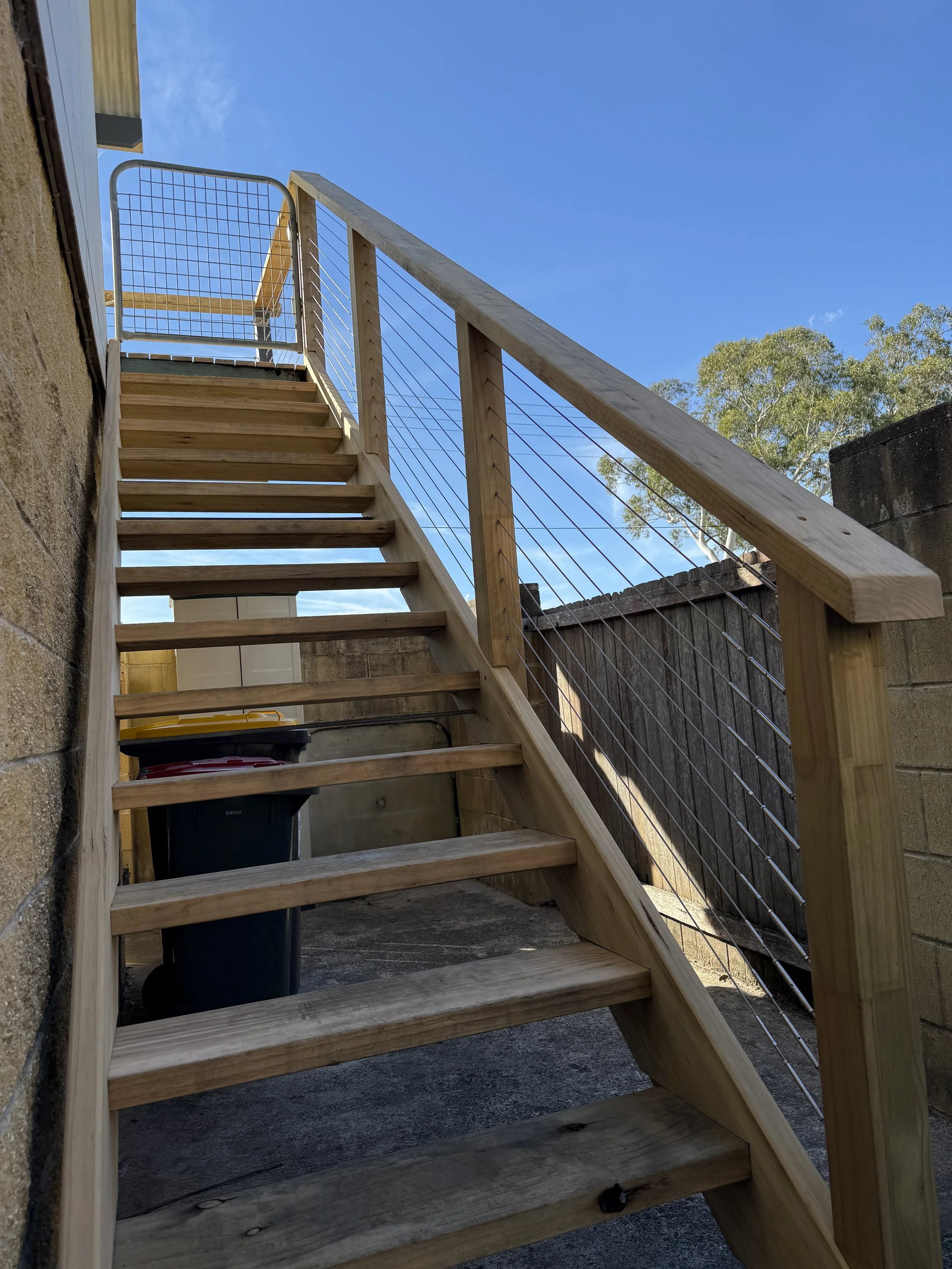 Wooden outdoor staircase with wire railing, leading up to a higher level, set against a blue sky with some trees in the background.