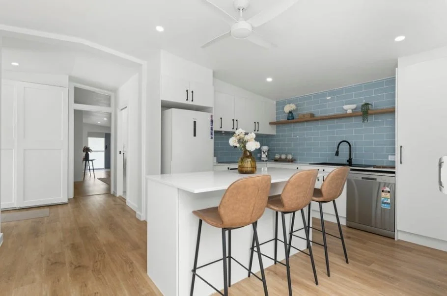 Modern kitchen with white cabinets, wooden shelves, blue tile backsplash, three brown bar stools, and a vase of flowers on the counter.