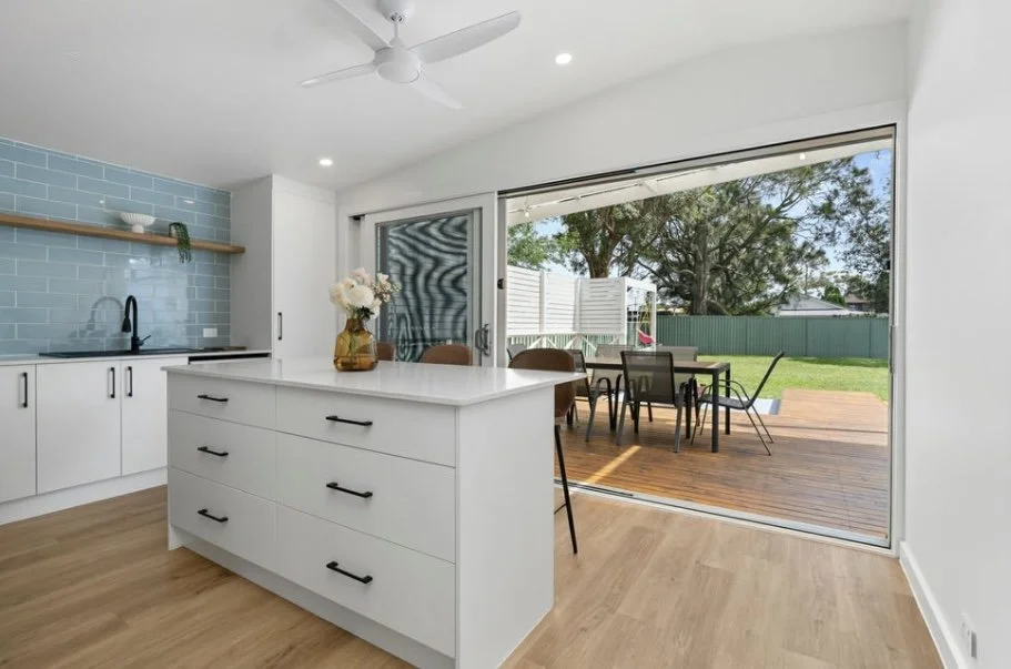 Bright kitchen with light wooden flooring, white cabinets, and a sliding glass door leading to outdoor patio and backyard with green lawn and trees.