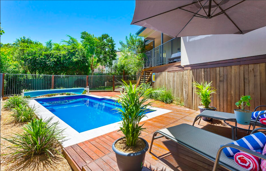 Backyard pool area with wooden deck, lounge chairs, and umbrella, surrounded by green plants and trees.