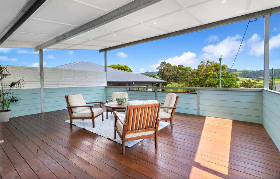 Outdoor balcony with wooden floor, four outdoor chairs and a round table, potted plant, and scenic view of trees and hills under a partly cloudy sky.
