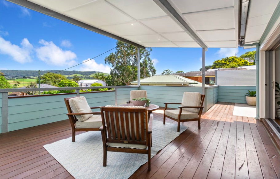 Outdoor balcony with wooden floor, seating area with four chairs and a round table, light blue railing, potted plant, scenic view of trees and hills under a partly cloudy sky.