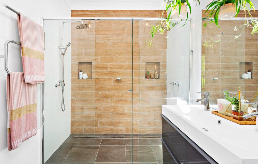 Modern bathroom with glass-enclosed shower, wooden wall tiles, white sink with black cabinet, pink and yellow towels, indoor plants, and vaulted glass window.