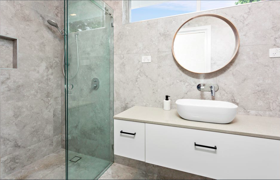 Modern bathroom with a glass-enclosed shower, a white vanity with a vessel sink, a round mirror, and a window
