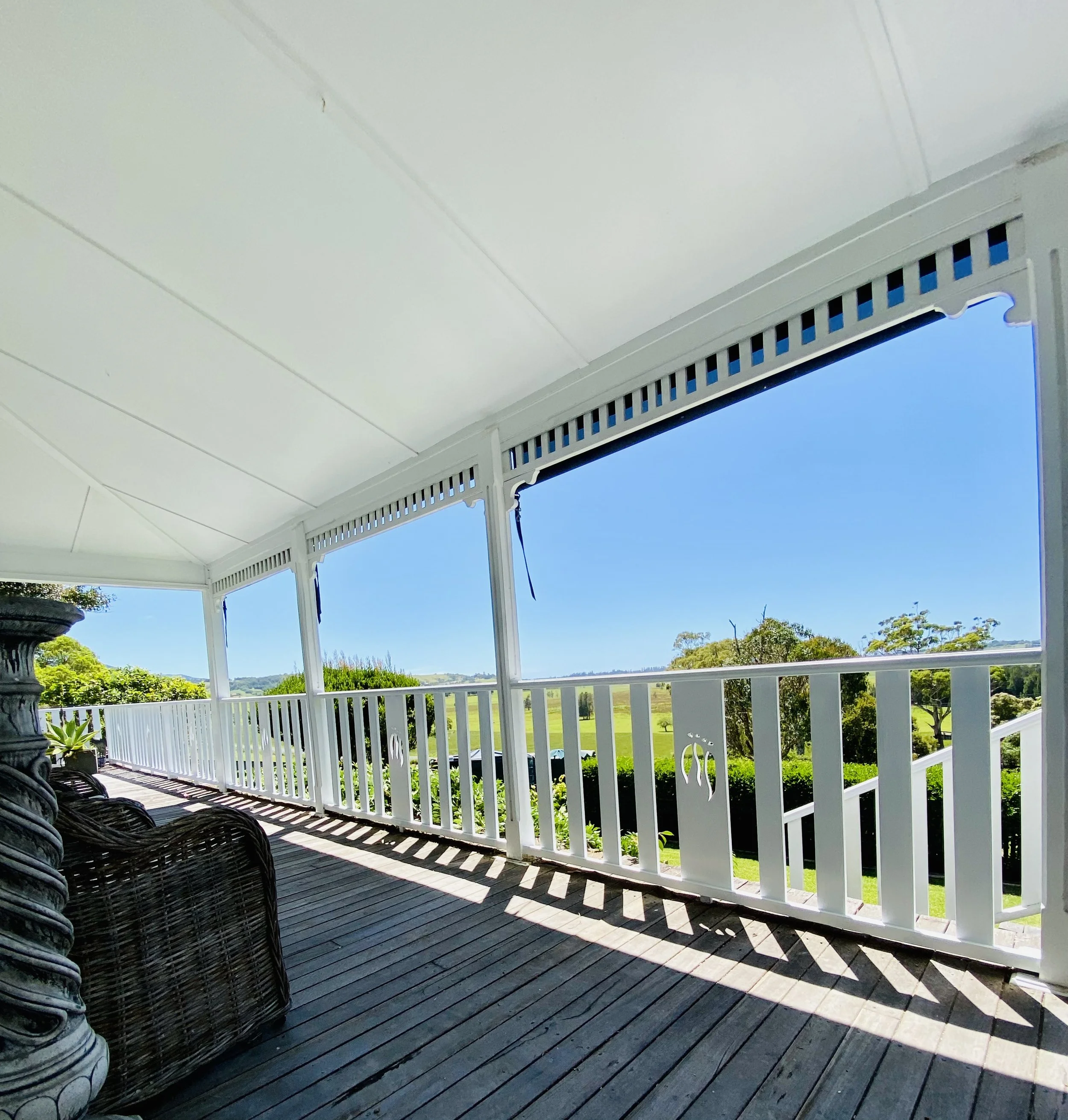 Covered porch of a house with white railing and siding, wicker chairs, and views of green fields and trees in the distance on a sunny day.