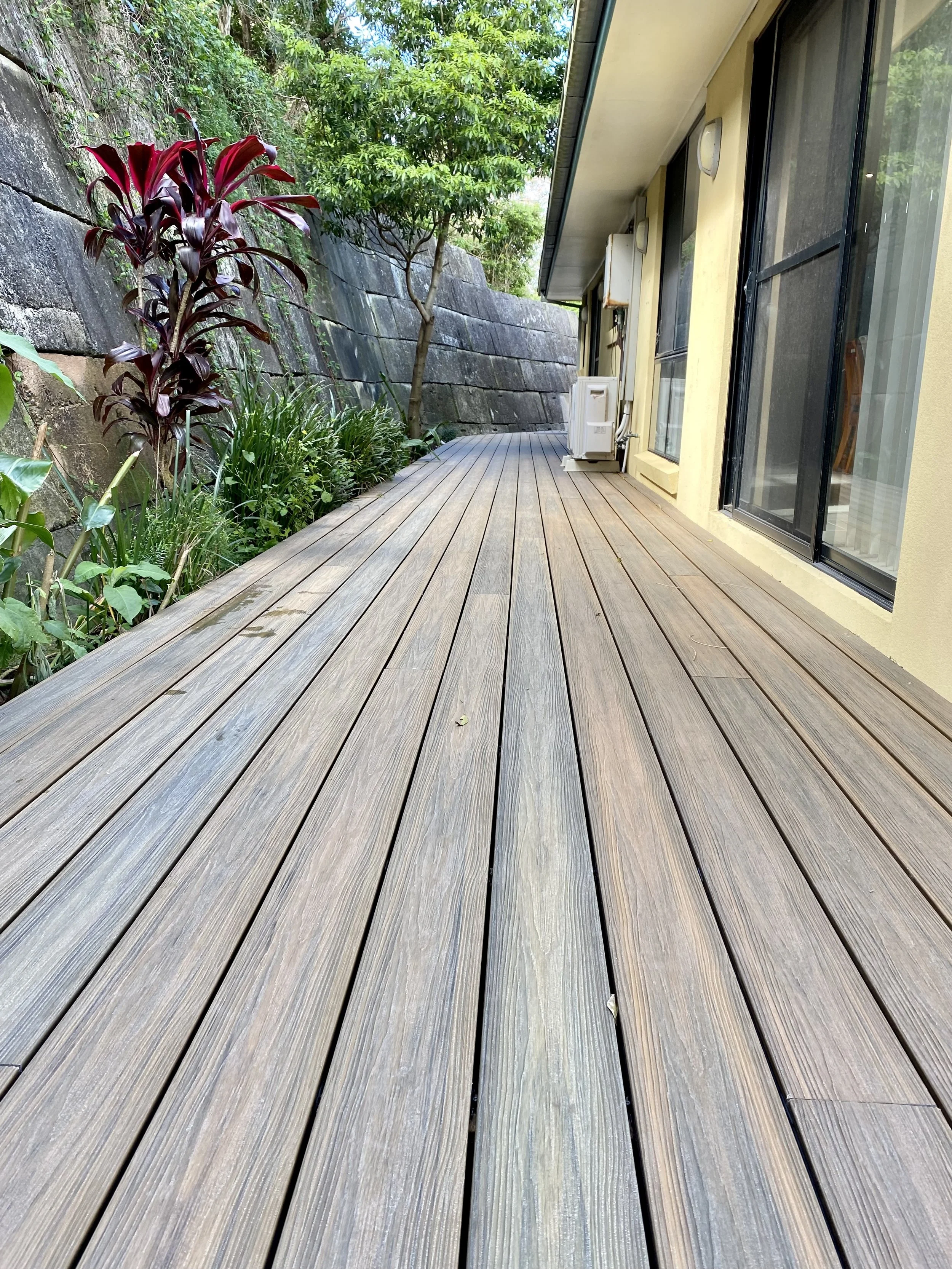 A wooden deck with outdoor air conditioning units beside a yellow house, surrounded by plants and a stone retaining wall with greenery, trees, and bushes in the background.