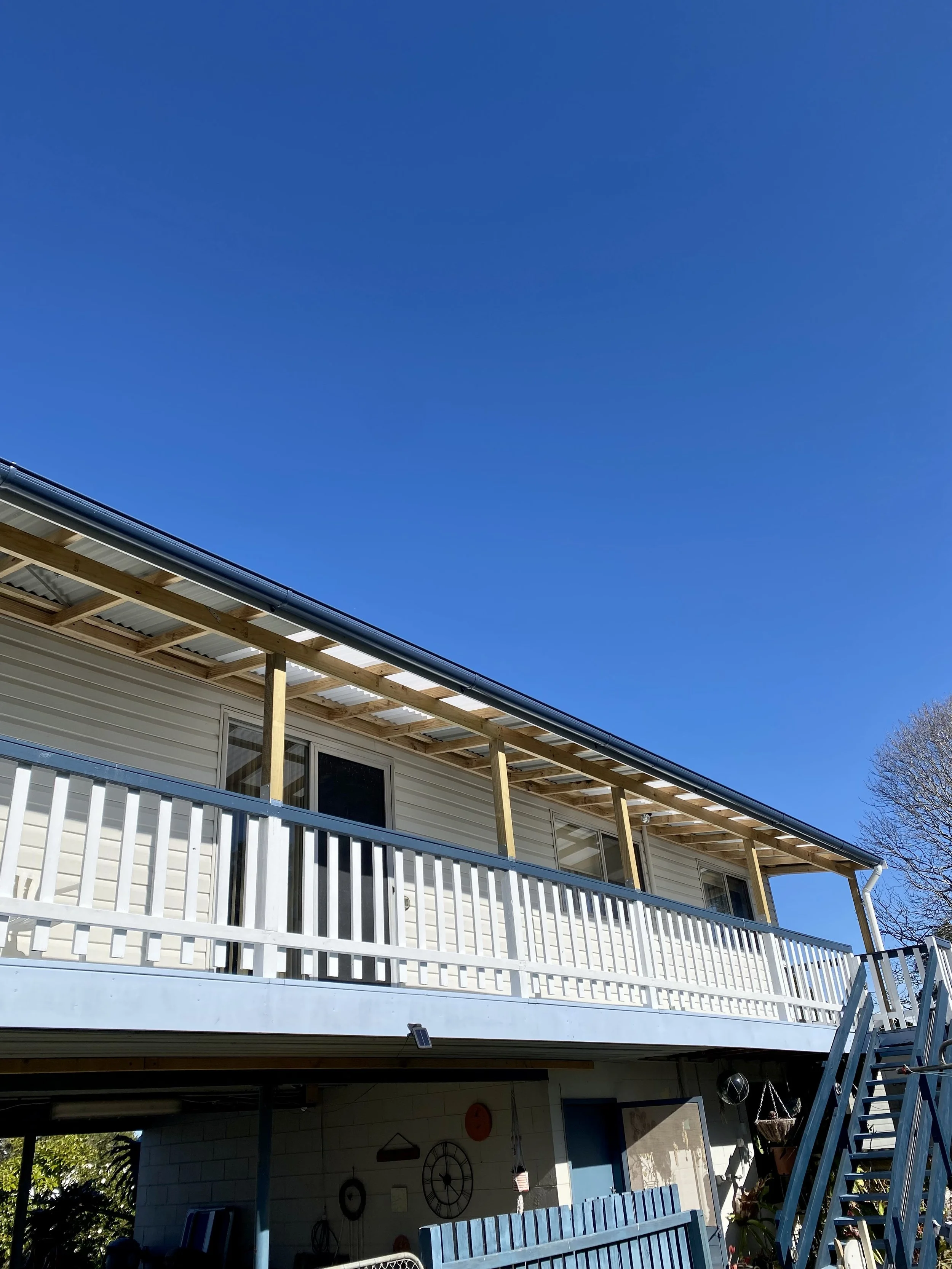 Backyard deck under construction with wooden railing and stairs, part of a house with a beige exterior, and a clear blue sky.
