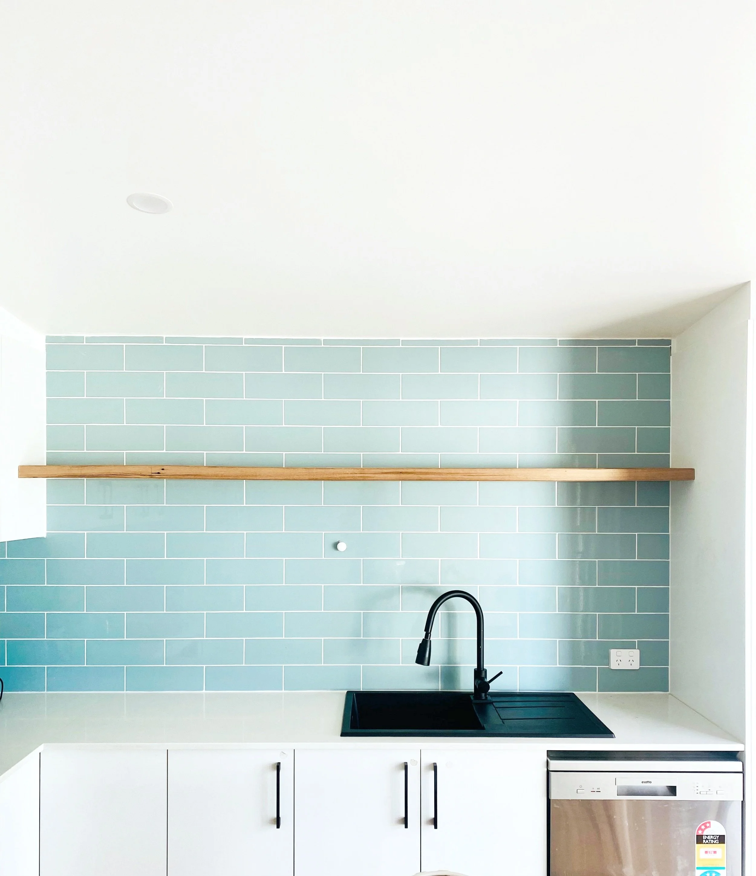 Modern kitchen with light blue subway tile backsplash, black faucet, white cabinetry, wooden shelf, and stainless steel dishwasher.