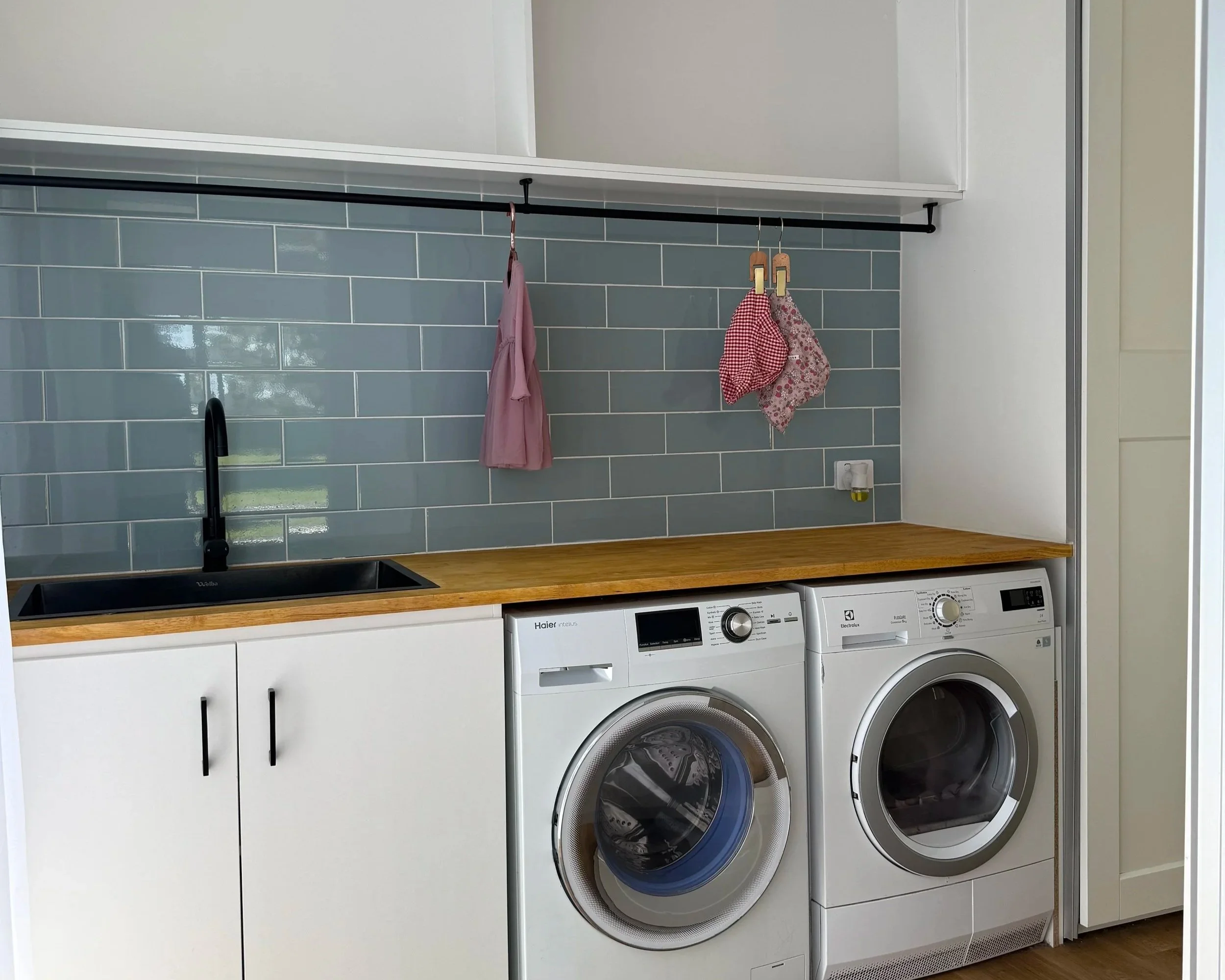 A laundry room with a wooden countertop above white washer and dryer units, a grey tiled backsplash, a black hanging rod, and two hanging cloths or towels.