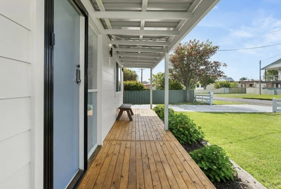 View of a wooden porch with a bench, white house siding, and a green lawn with trees and a street in the background.