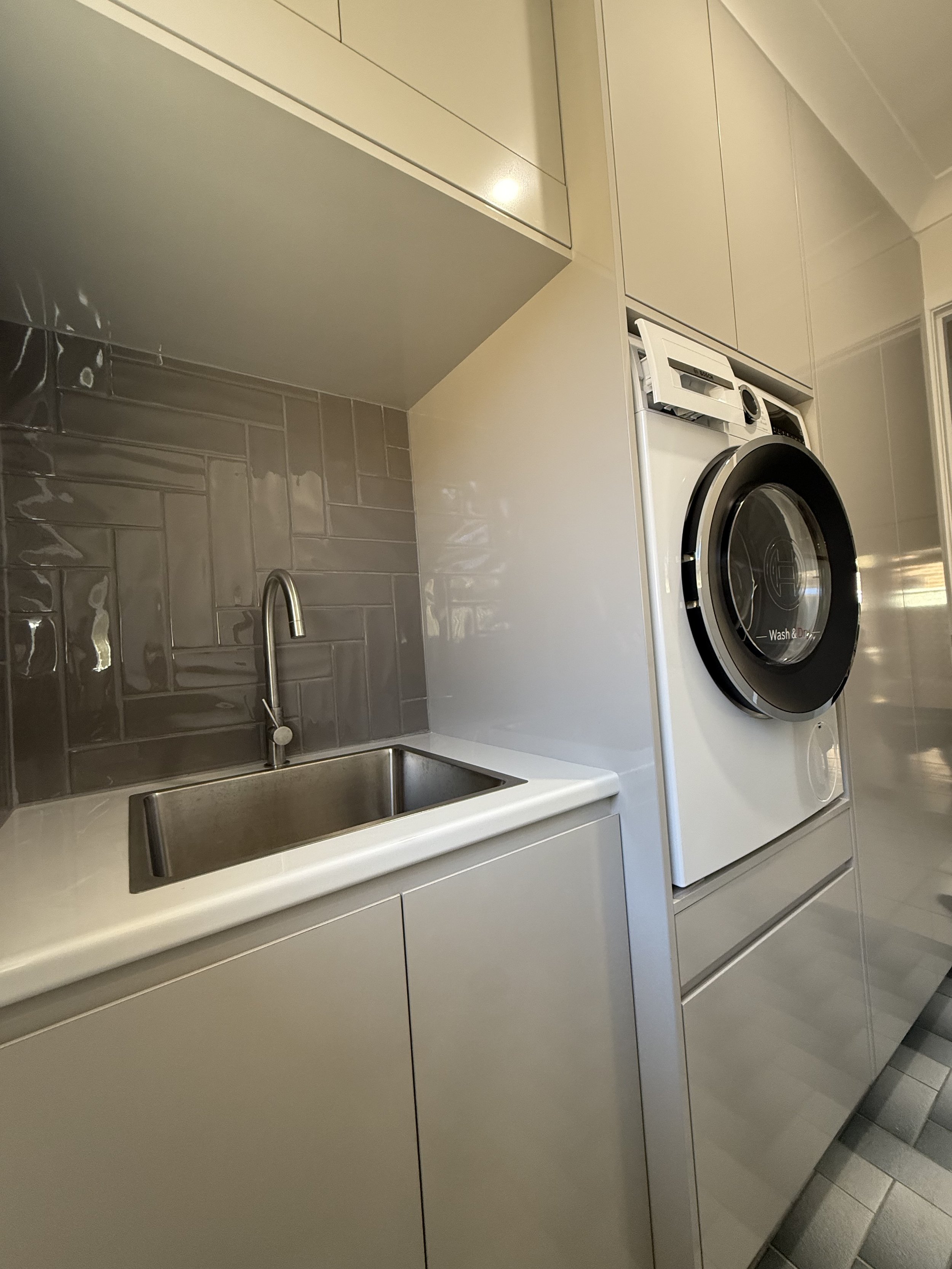 A modern laundry room with beige cabinets, a black and white front-loading washing machine, a white countertop, a stainless steel sink, and gray tile flooring.