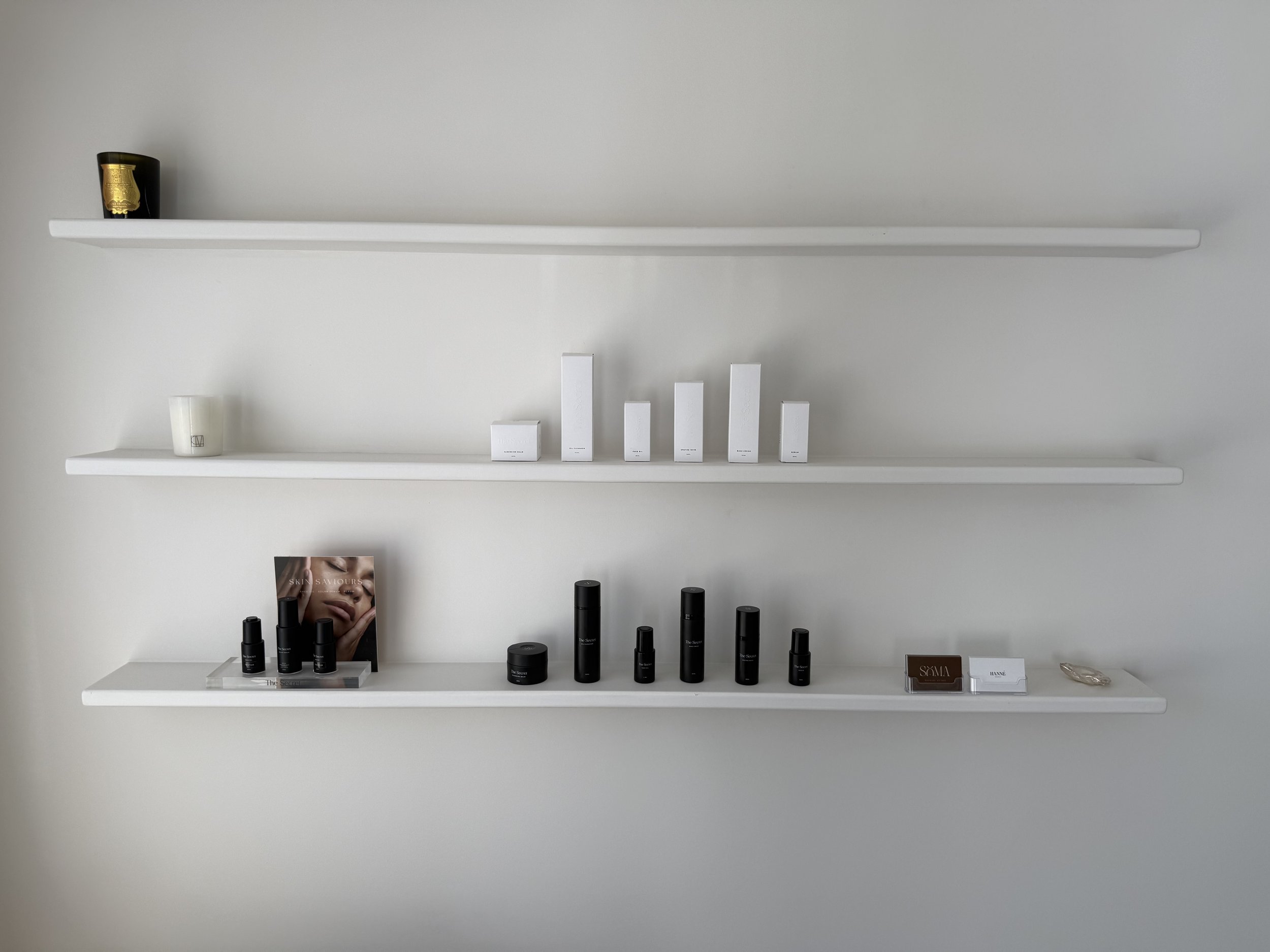 Three white shelves with skincare and cosmetic products, some in black bottles, a skincare magazine, and a small sign on a clean white wall.