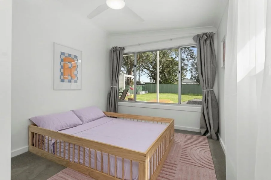 A bedroom featuring a wooden bed frame with lavender bedding, gray curtains on a large window, a pink rug, and artwork on the white walls.
