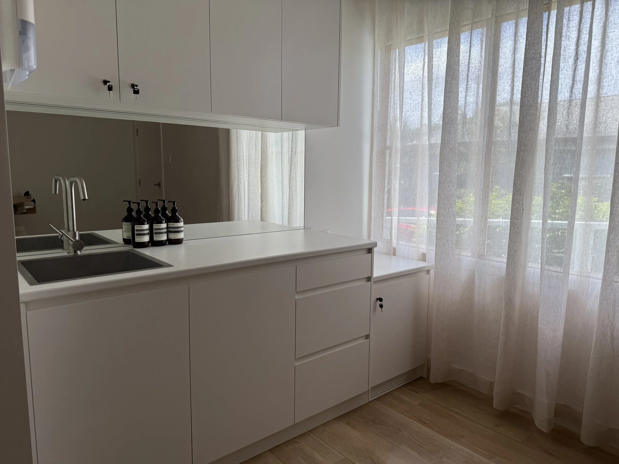 A modern white kitchen with a countertop, a sink, four bottles of soap or lotion on the counter, white cabinets, a wooden floor, and sheer curtains on large windows allowing natural light.