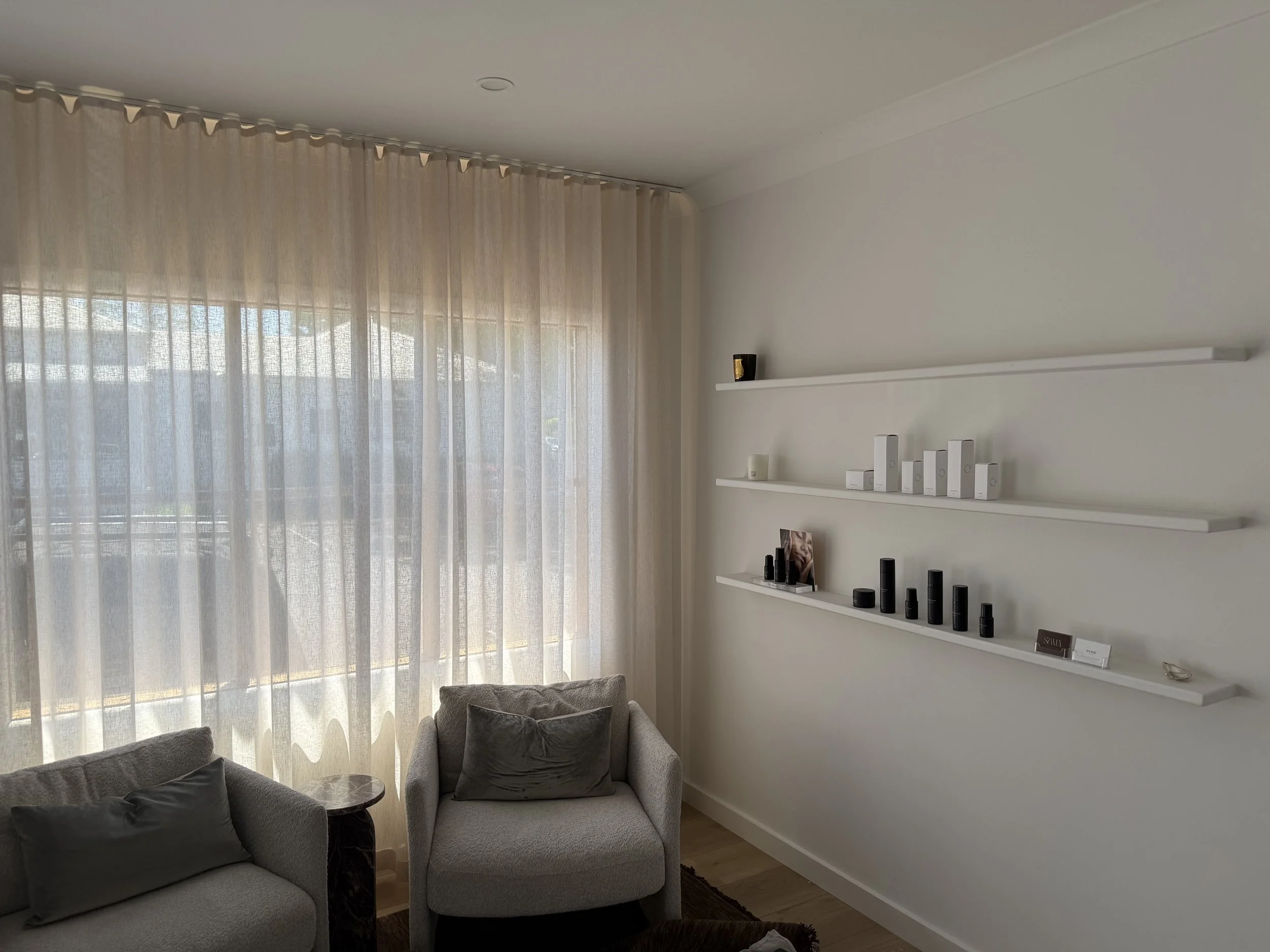 Living room corner with a light gray armchair and a gray sofa, curtains covering large windows, and minimalist white wall shelves with decorative items.
