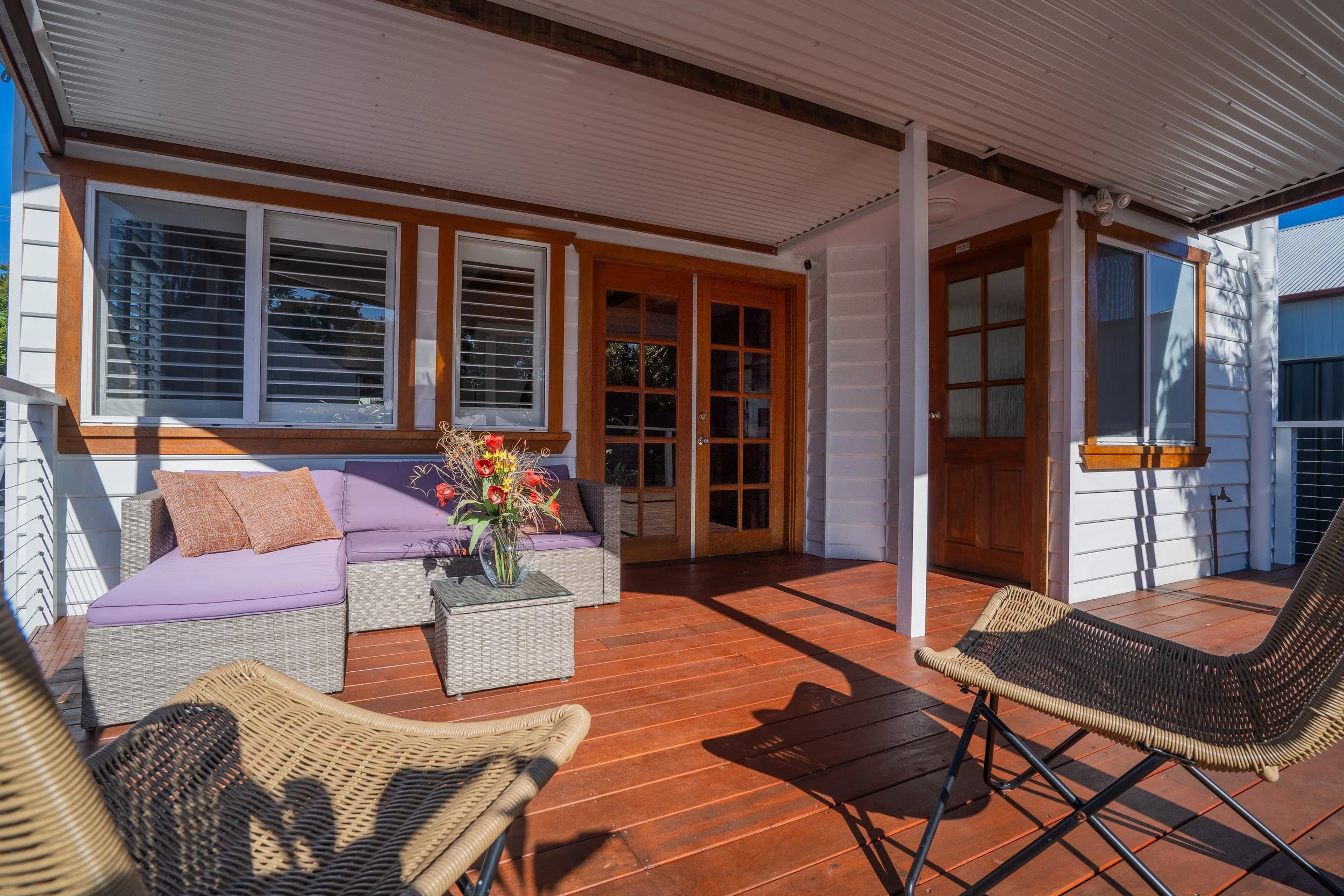 A covered outdoor patio with rattan furniture, including a purple cushioned sofa and a small coffee table with a flower arrangement. There are additional wicker chairs and black metal chairs on a wooden deck, with large windows and glass-paneled door