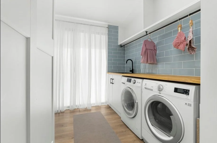 A laundry room with white cabinets, a wooden countertop, and a blue tiled backsplash. There are two front-loading washing machines, a small black faucet, and pink baby clothes hanging on a rod.
