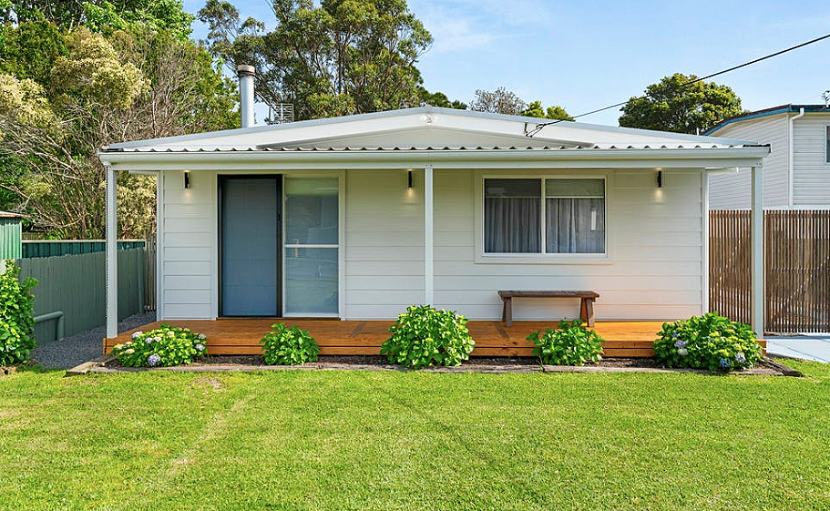 Small white house with a porch, wooden steps, and a bench. Green shrubs and grass in front, trees behind, and neighboring houses visible.