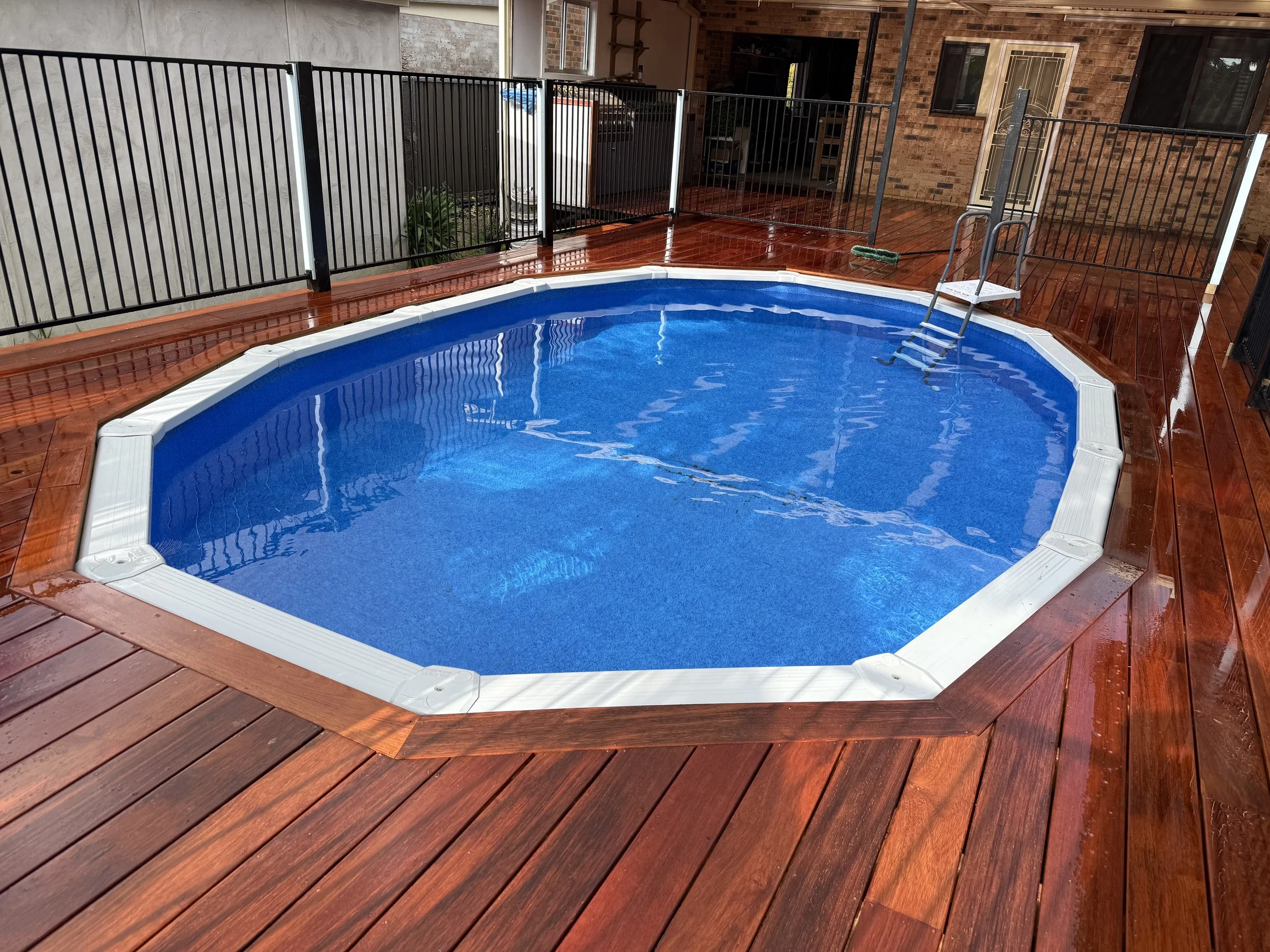 Backyard with a small above-ground swimming pool with blue water, wooden deck, black metal safety fence, a ladder, and behind it a brick wall house with a door and window.