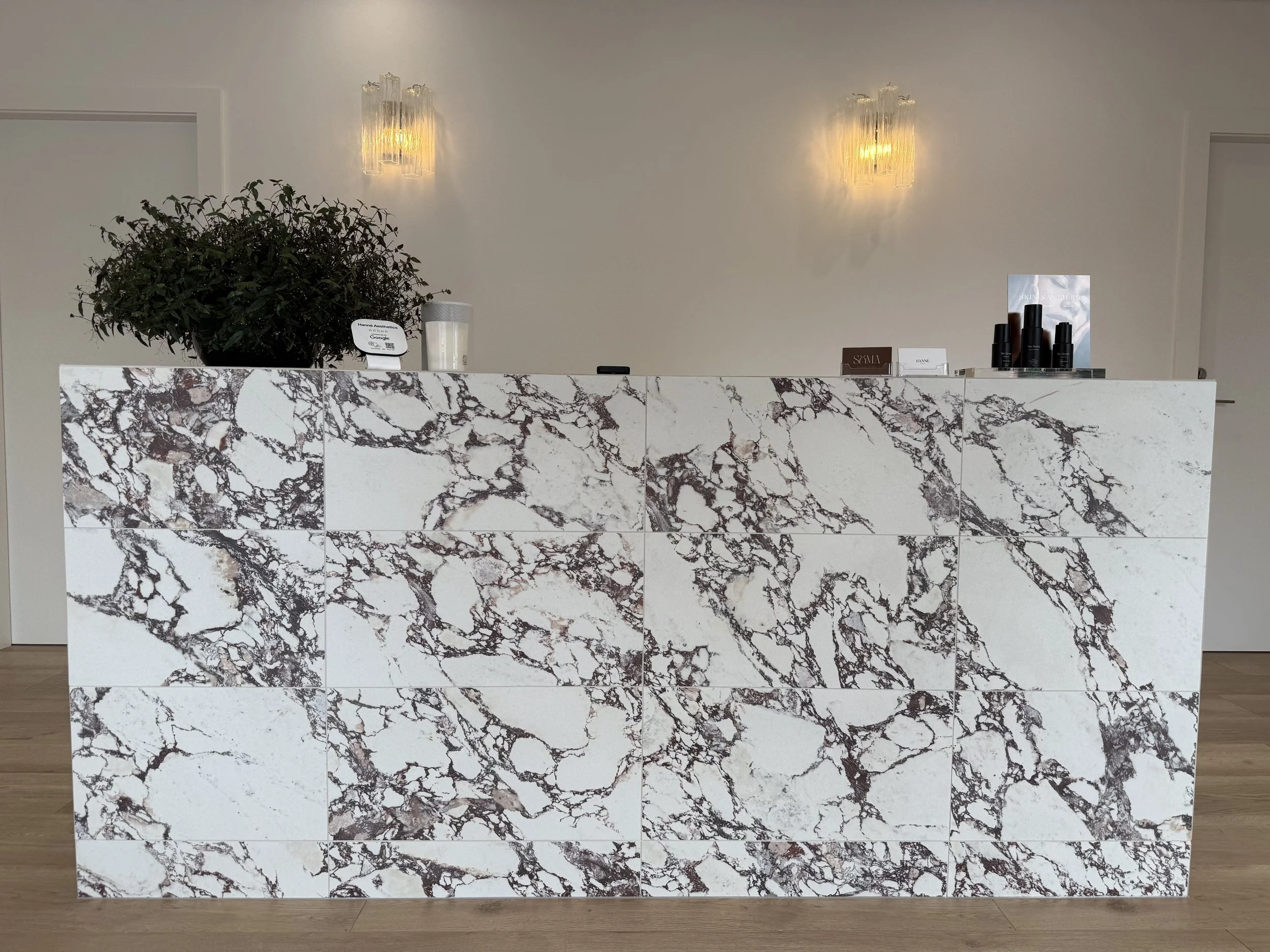 A marble counter with a potted plant, a candle, and black bottles for skincare products. The wall behind has two decorative wall sconces and some small signs. The floor is wooden.