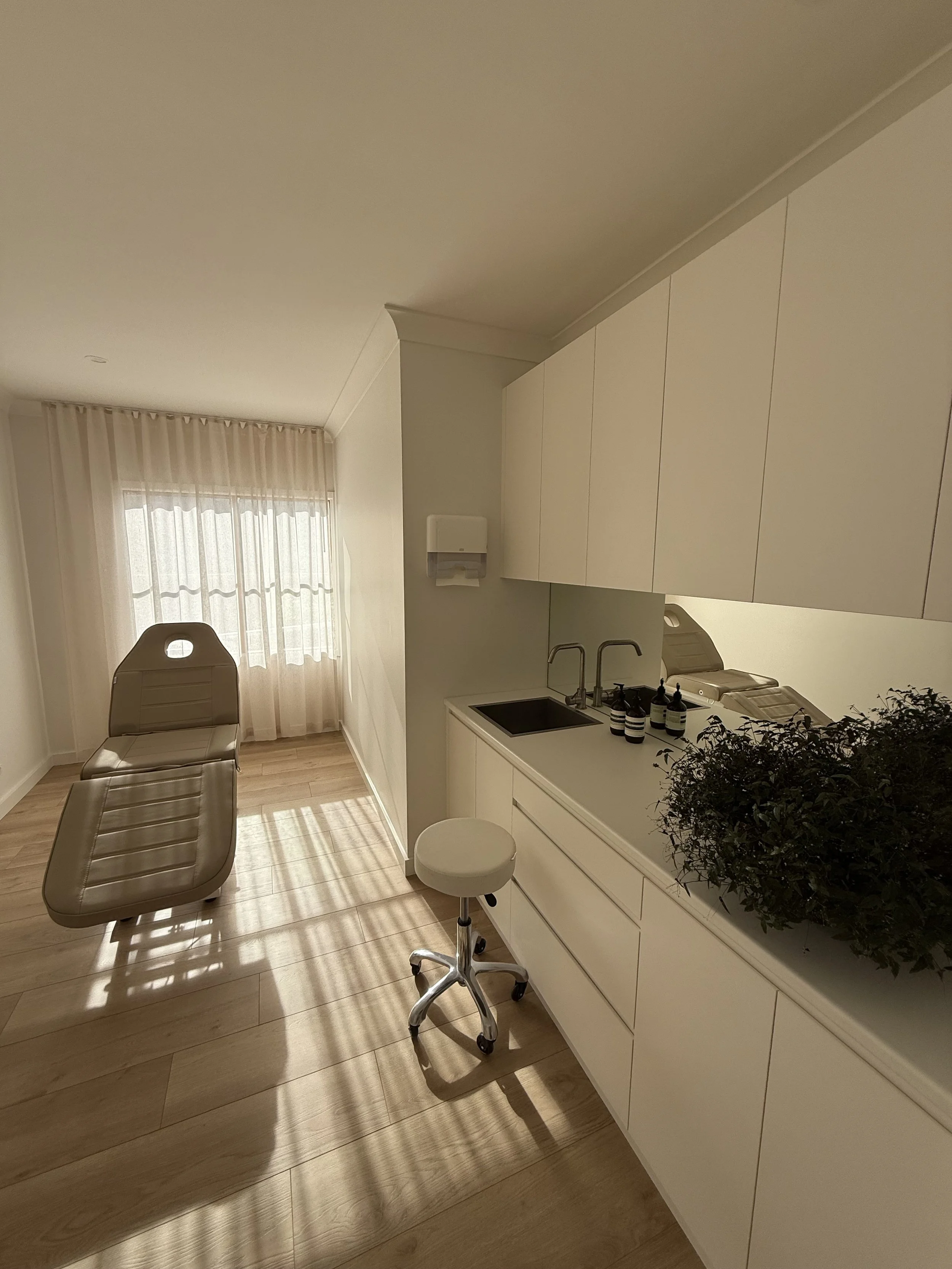 A room with beige chairs, white cabinets, a small sink, soap/disinfectant bottles, a rolling stool, a window with sheer curtains, and a potted plant, illuminated by sunlight.