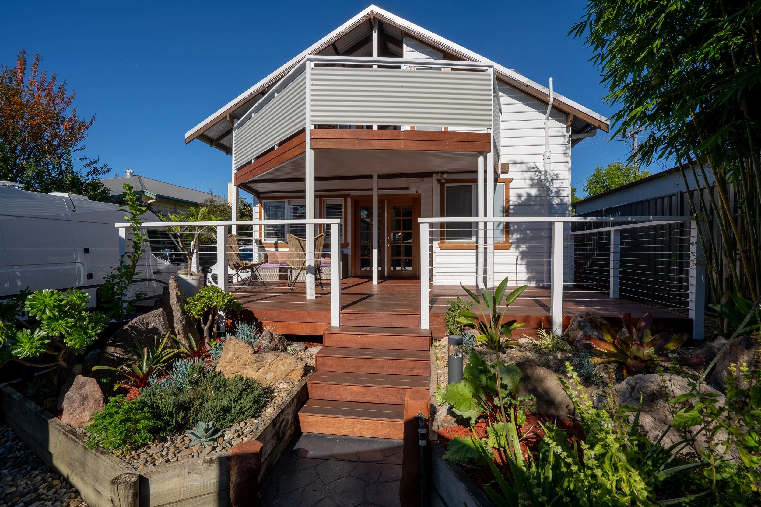 A two-story house with a wooden deck, outdoor furniture, and surrounding plants and rocks in a garden.