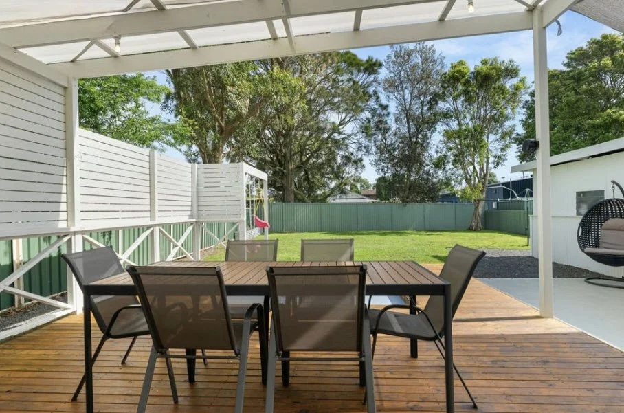 Covered backyard patio with a wooden table and six chairs, fenced yard with grass and trees, and a children's swing set in the background.