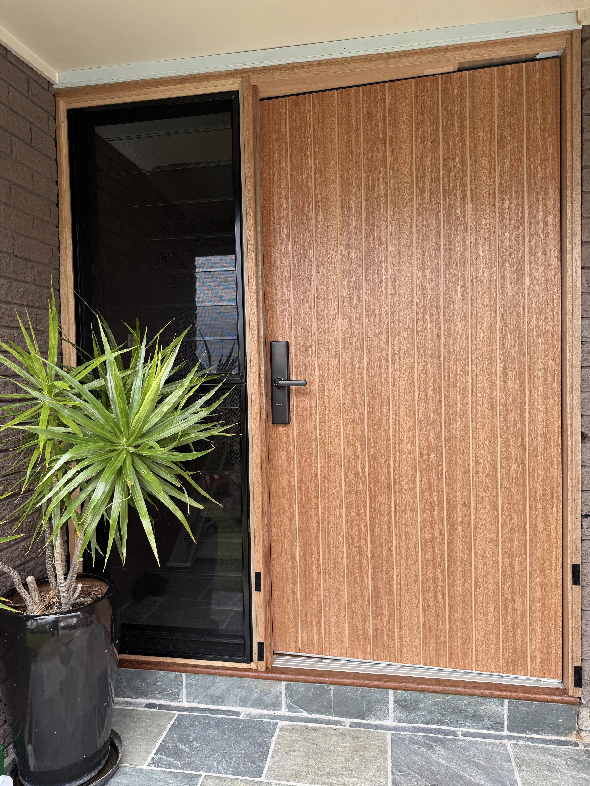 A front door with a sliding wooden door and a glass panel door on the left, next to a large potted plant with green leafy foliage, on a tiled floor.