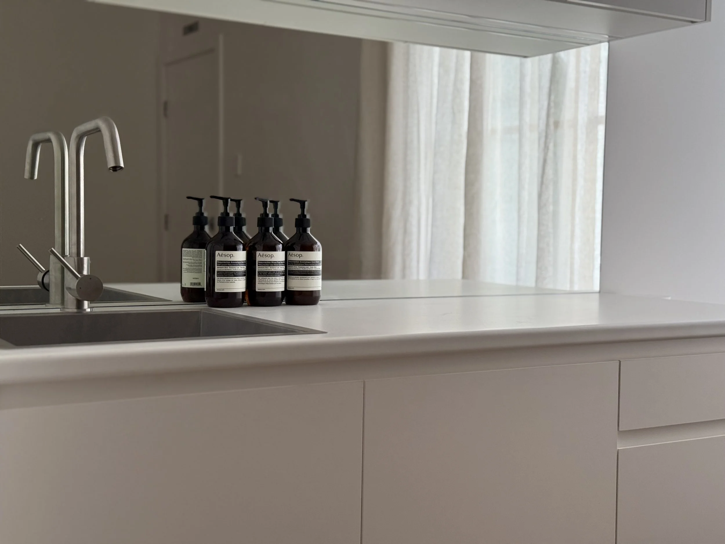 A modern kitchen featuring a white countertop, a stainless steel faucet, and four brown bottles of Aesop hand soap, with a window and white curtains in the background.