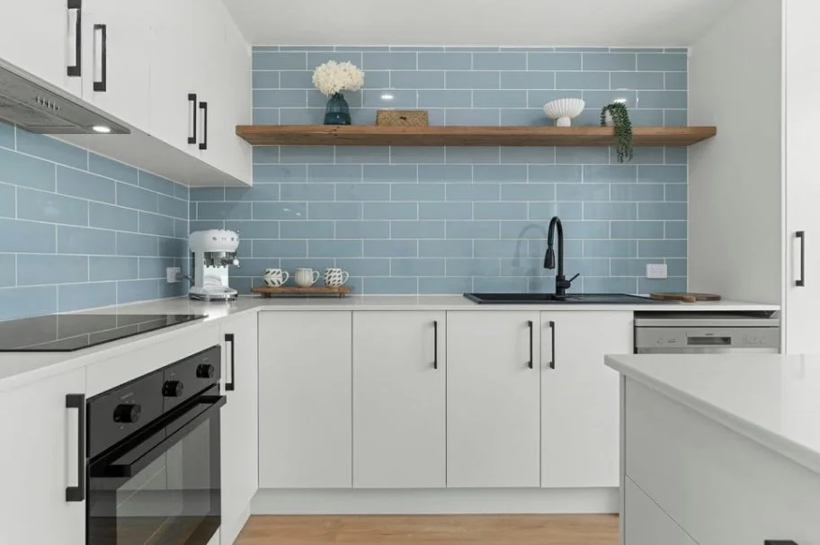Modern kitchen with white cabinets, black handles, a black backsplash, and a light blue tile wall. Features include a built-in oven, a coffee machine, some cups, a wooden shelf with decorative items, a black faucet, and a dishwasher.
