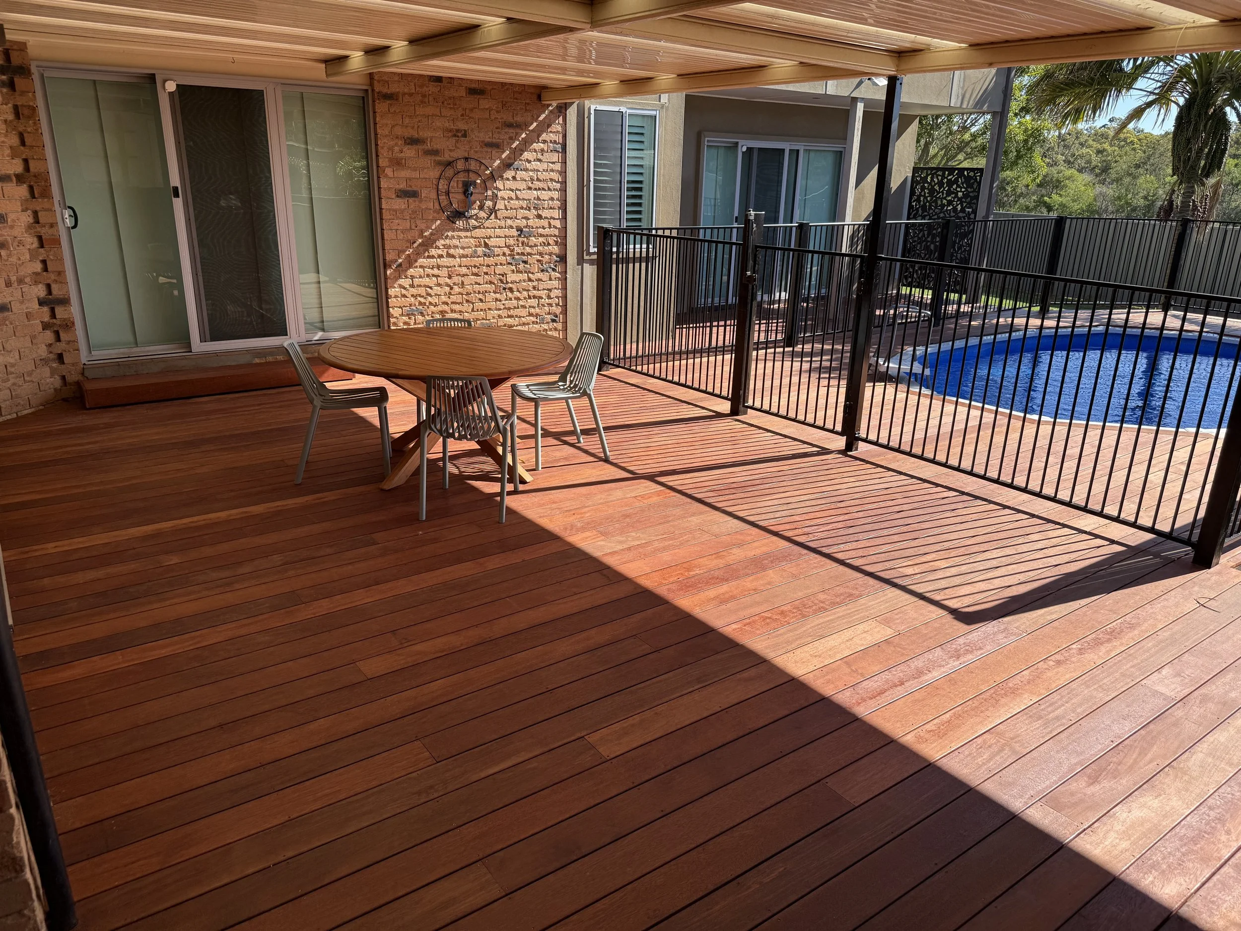 Covered wooden patio with outdoor dining table and chairs, brick wall, sliding glass door, swimming pool, and surrounding trees.