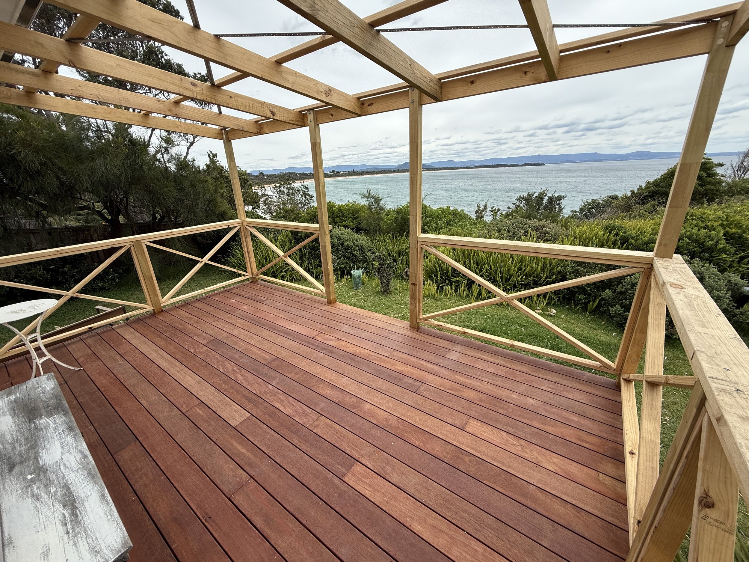 The image shows an unfinished wooden balcony overlooking trees, a body of water, and distant mountains on a cloudy day.